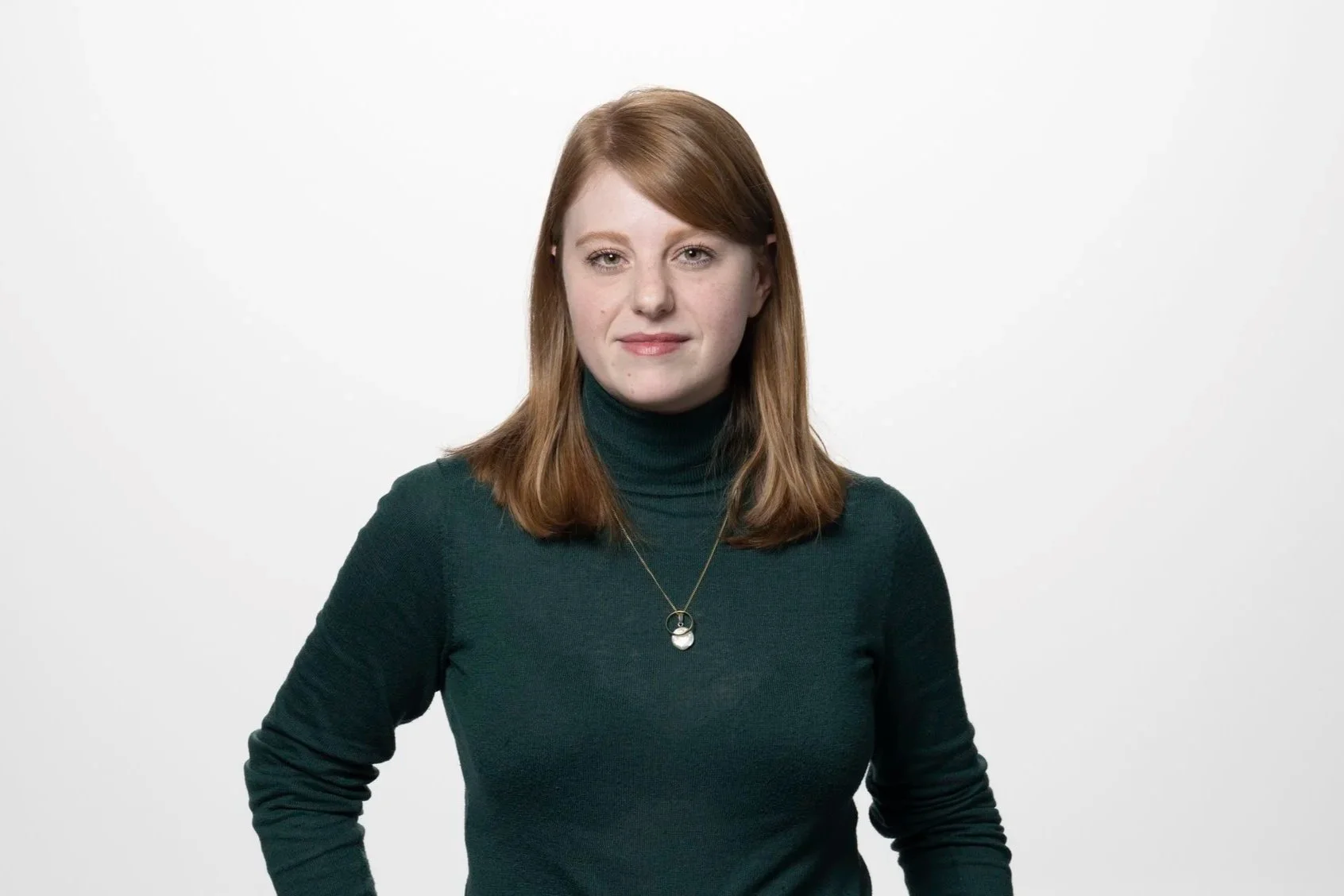 Eine junge Frau mit rotem Haar, trägt einen dunkelgrünen Rollkragenpullover und eine Halskette, steht vor einem weißen Hintergrund.