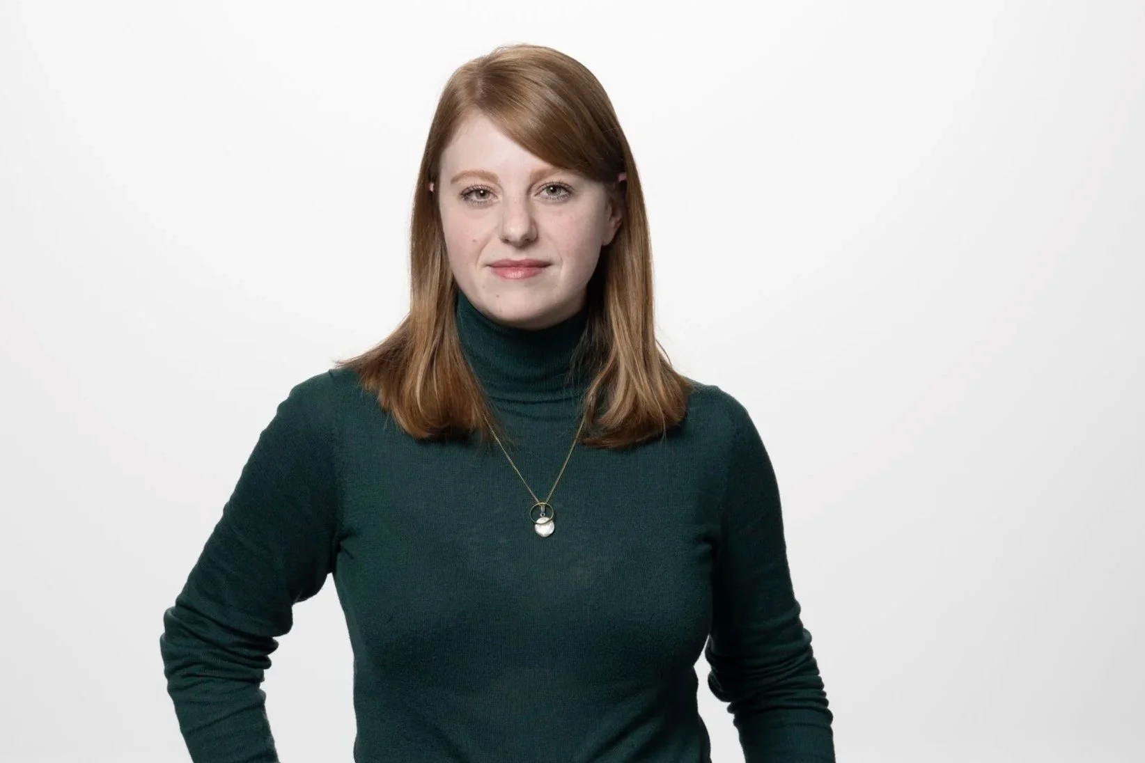 Junge Frau mit braunen, schulterlangen Haaren, trägt eine dunkelgrüne Rollkragenpullover und eine Halskette, vor weißem Hintergrund.