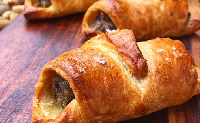 Close-up of two cooked sausage rolls with golden-brown pastry, resting on a wooden surface, with visible dark sausage filling inside.