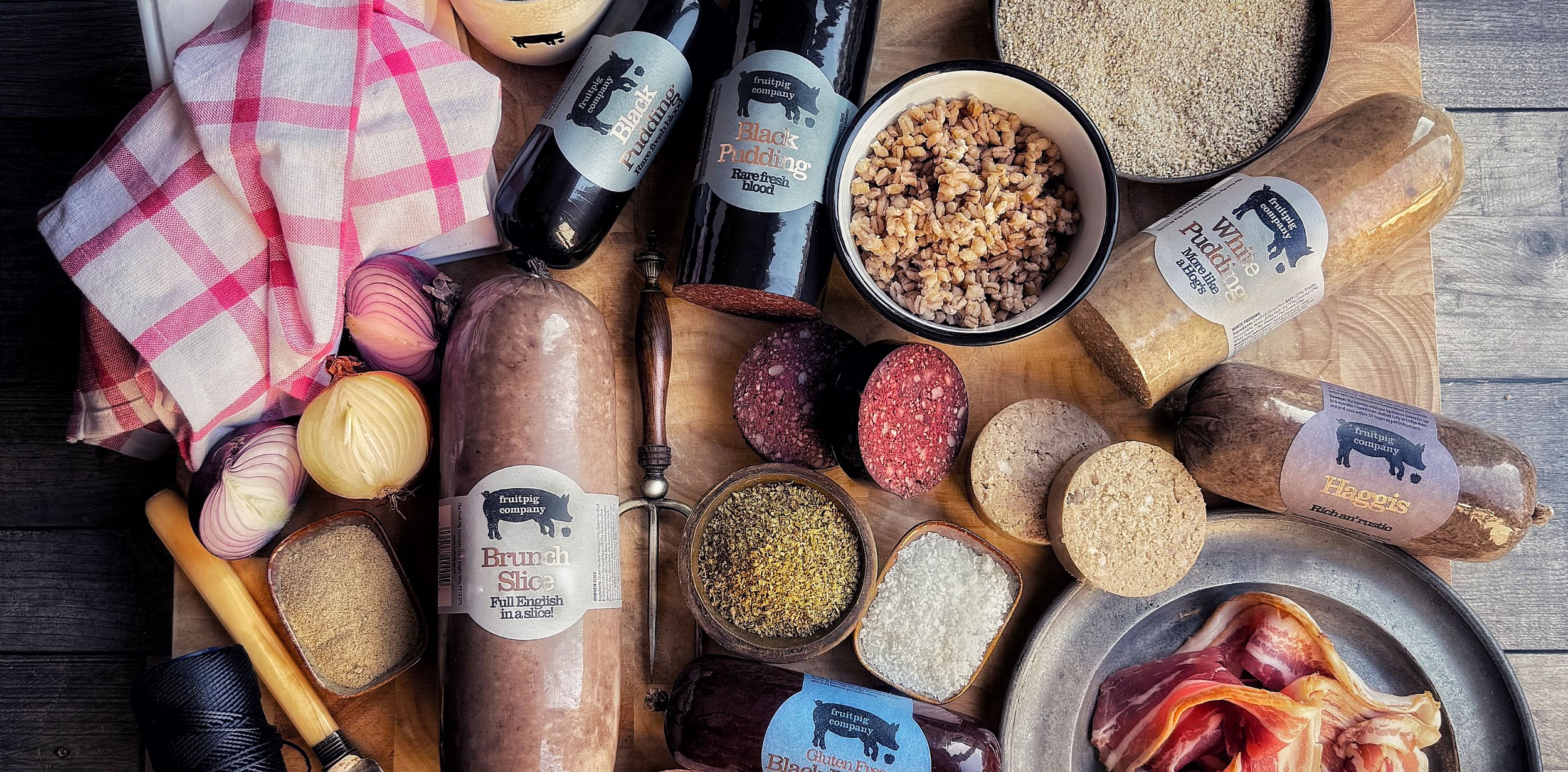 A wooden table filled with ingredients and Fruit Pig products, including black pudding, haggis, white pudding and spices.