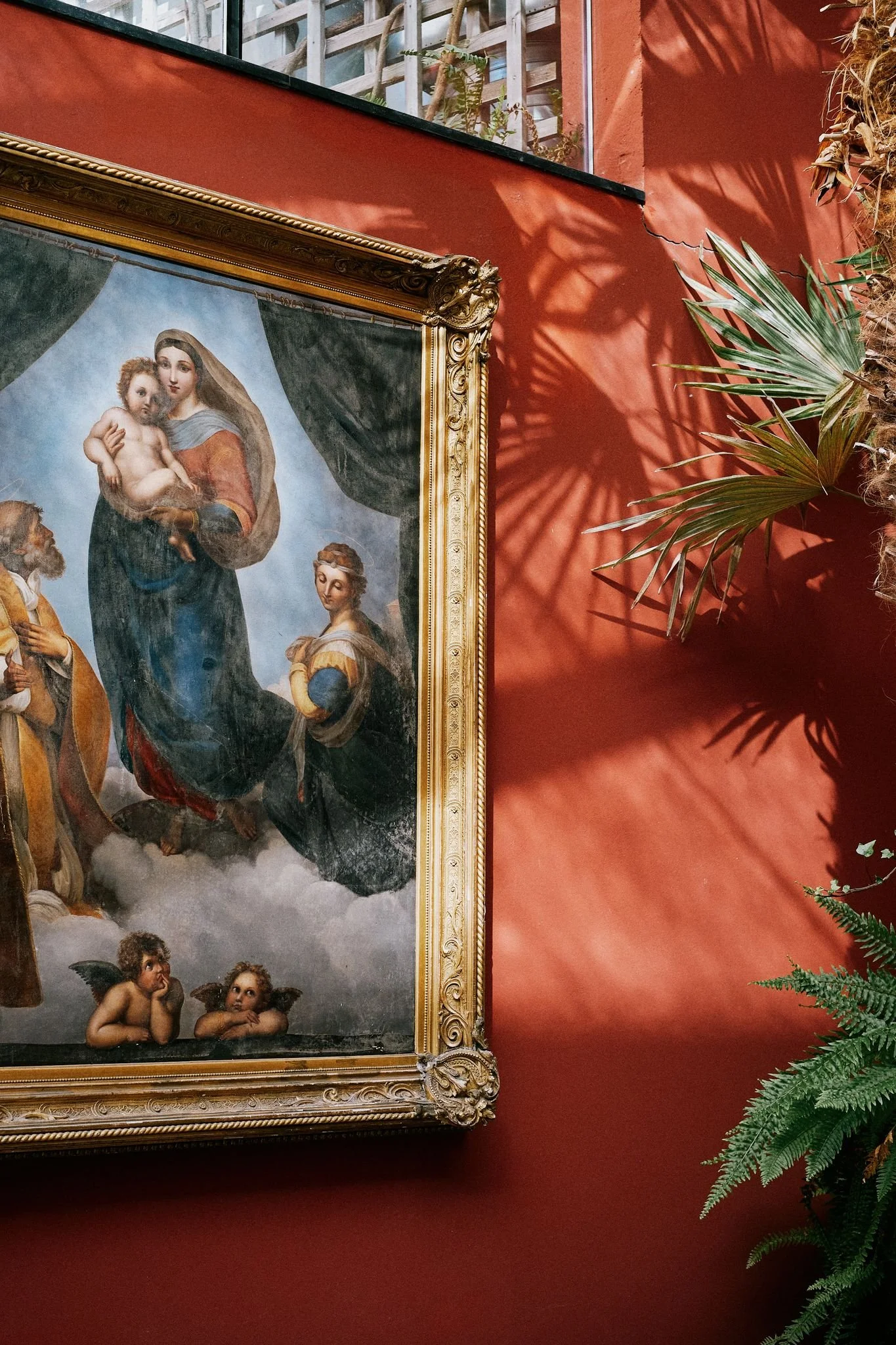 En målning av den heliga familjen med Jungfru Maria, Jesusbarnet och två änglar, omgiven av gröna växter på en röd vägg.