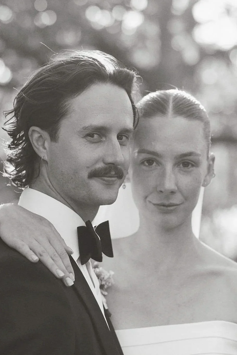Svartvitt foto av ett par på bröllop, mannen i tuxedo med svart fluga och kvinnan i vit klänning, de står nära varandra och tittar mot kameran.