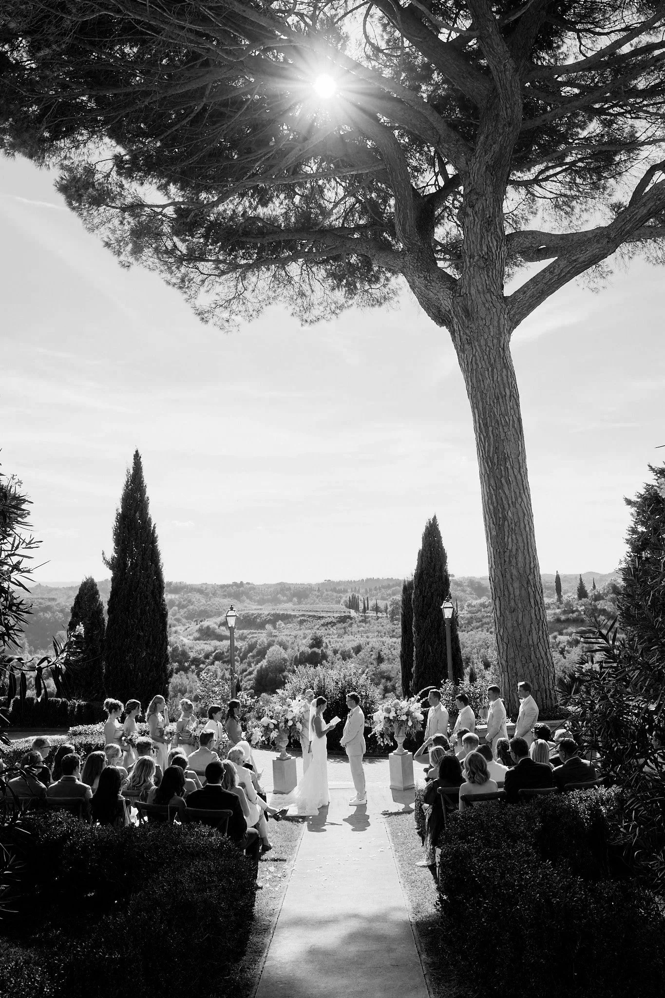 En utomhusbröllopsceremoni under en stor ek, med ett brudpar och vigselförrättare som står framför ett altare med blommor, omgiven av gäster.
