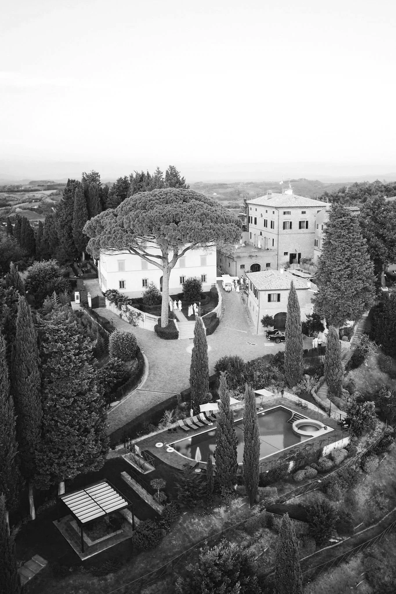 En svartvit bild av en villa med en stor liggande träd i mitten, omgiven av höga cypressträd och en pool i förgrunden.