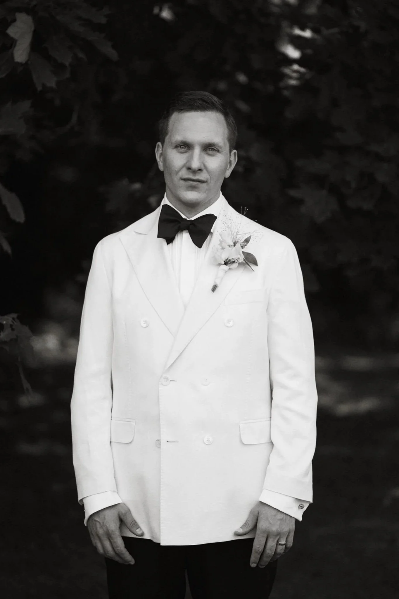 En man i en vit smoking med svart fluga och ett blomsterarrangemang på bröstet står mot en mörk bakgrund.