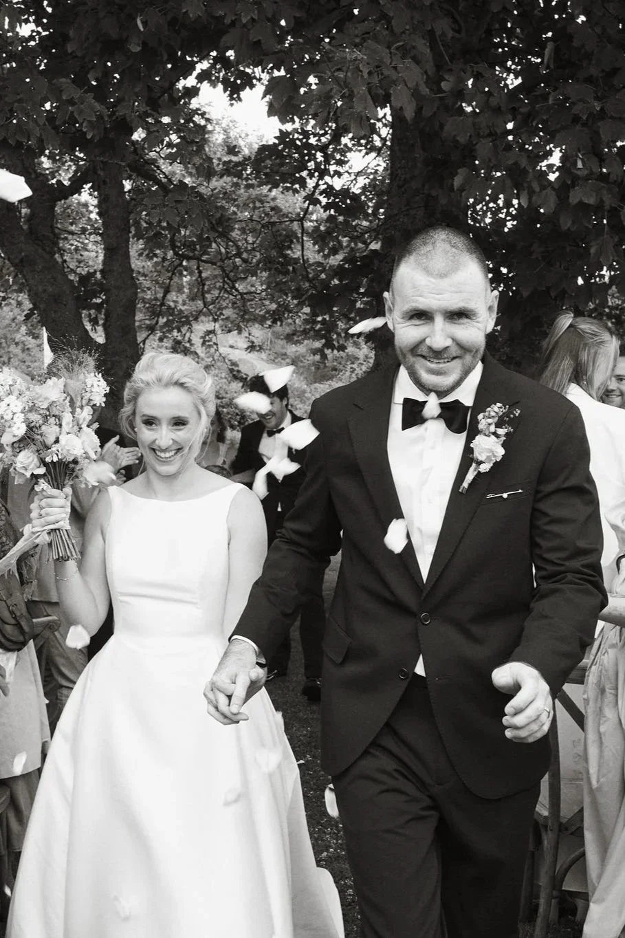 En brud och en brudgum går hand i hand på deras bröllopsfirande utomhus, med gäster runtomkring, blomsterarrangemang och träd i bakgrunden.