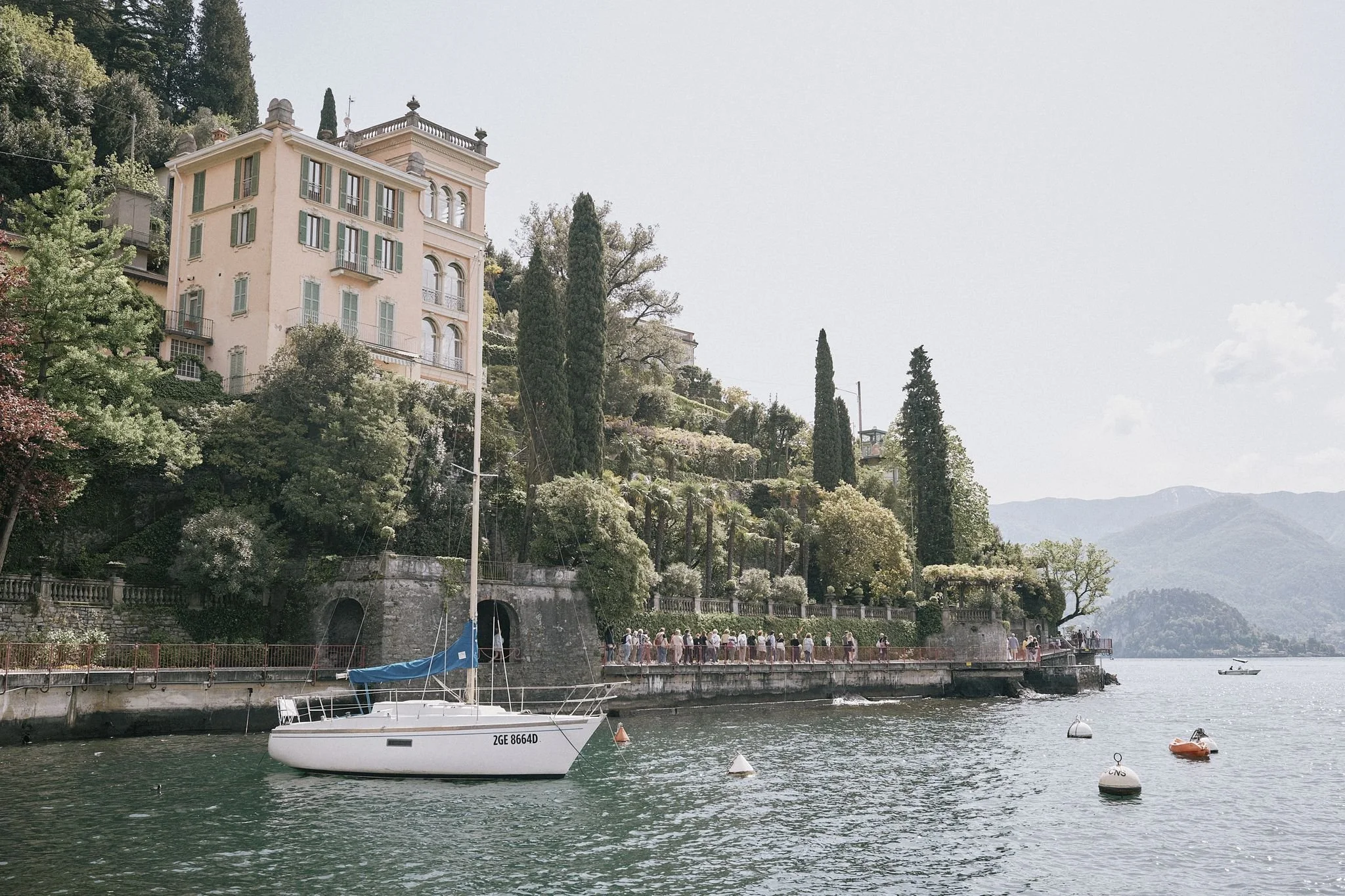 En vacker sjö med en vit segelbåt och flera mindre båtar, omgiven av gröna träd, ett gammaldags hus på klippor och fjäll i bakgrunden.