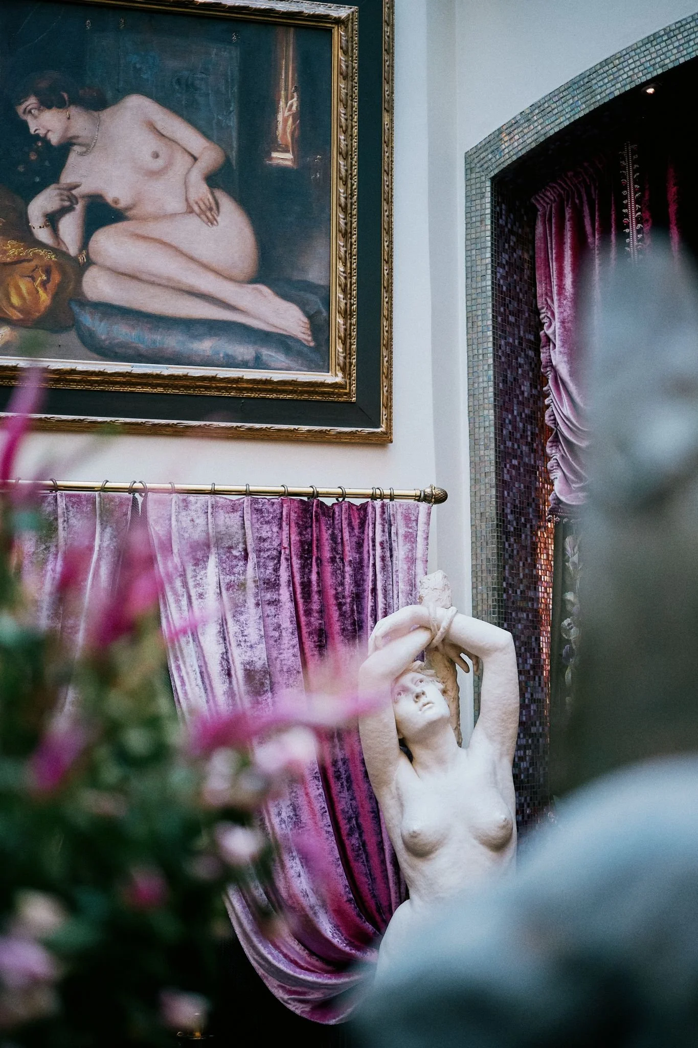 En konstnärlig bild av ett galleri eller museum med en målning av en naken kvinna på väggen, en skulptur av en naken kvinna i förgrunden, och röda sammetstapeter eller draperier. Det är ett konstnärligt och klassiskt inrett utrymme.