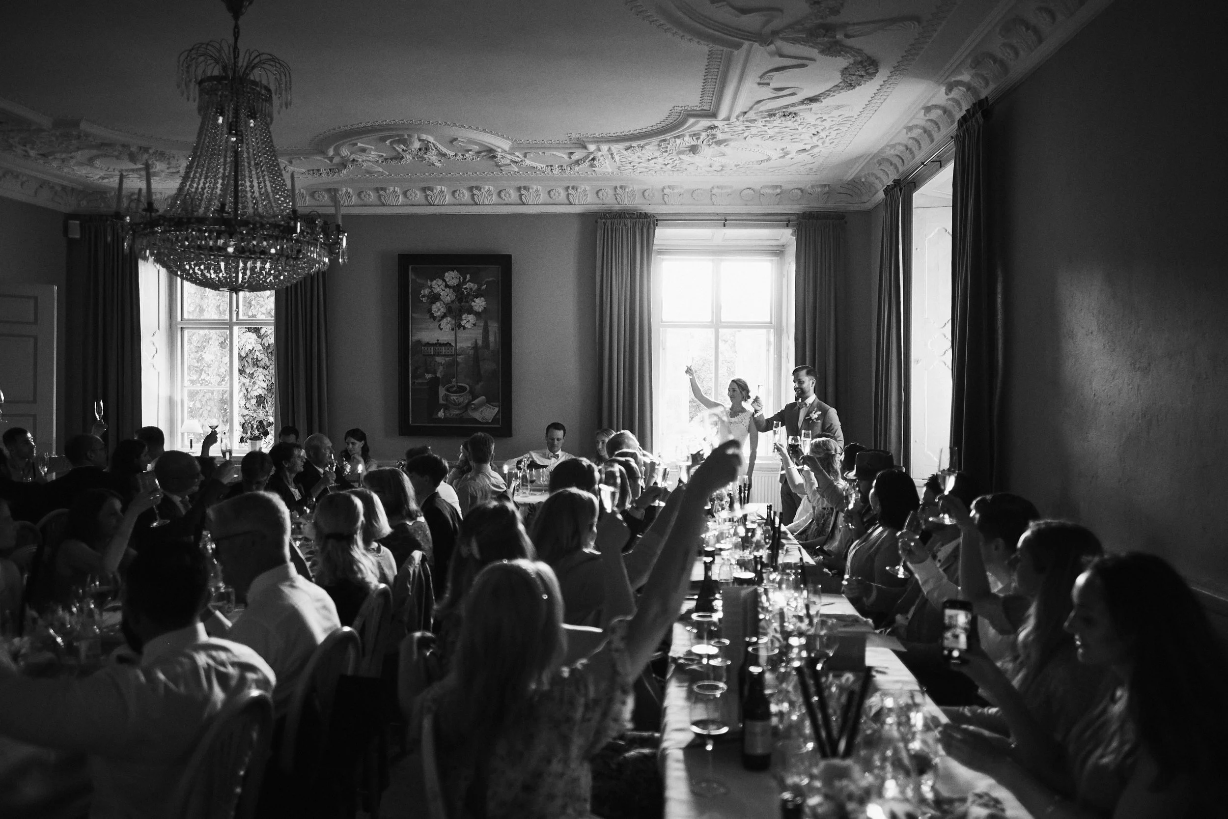 Svartvitt foto av ett festligt sällskap som samlats runt ett långbord i ett elegant rum. Personerna är i samtal och många håller i glas, några reser sig eller tar bilder, ett par står vid ett fönster och vinkar, taket är utsmyckat med en stor kristal