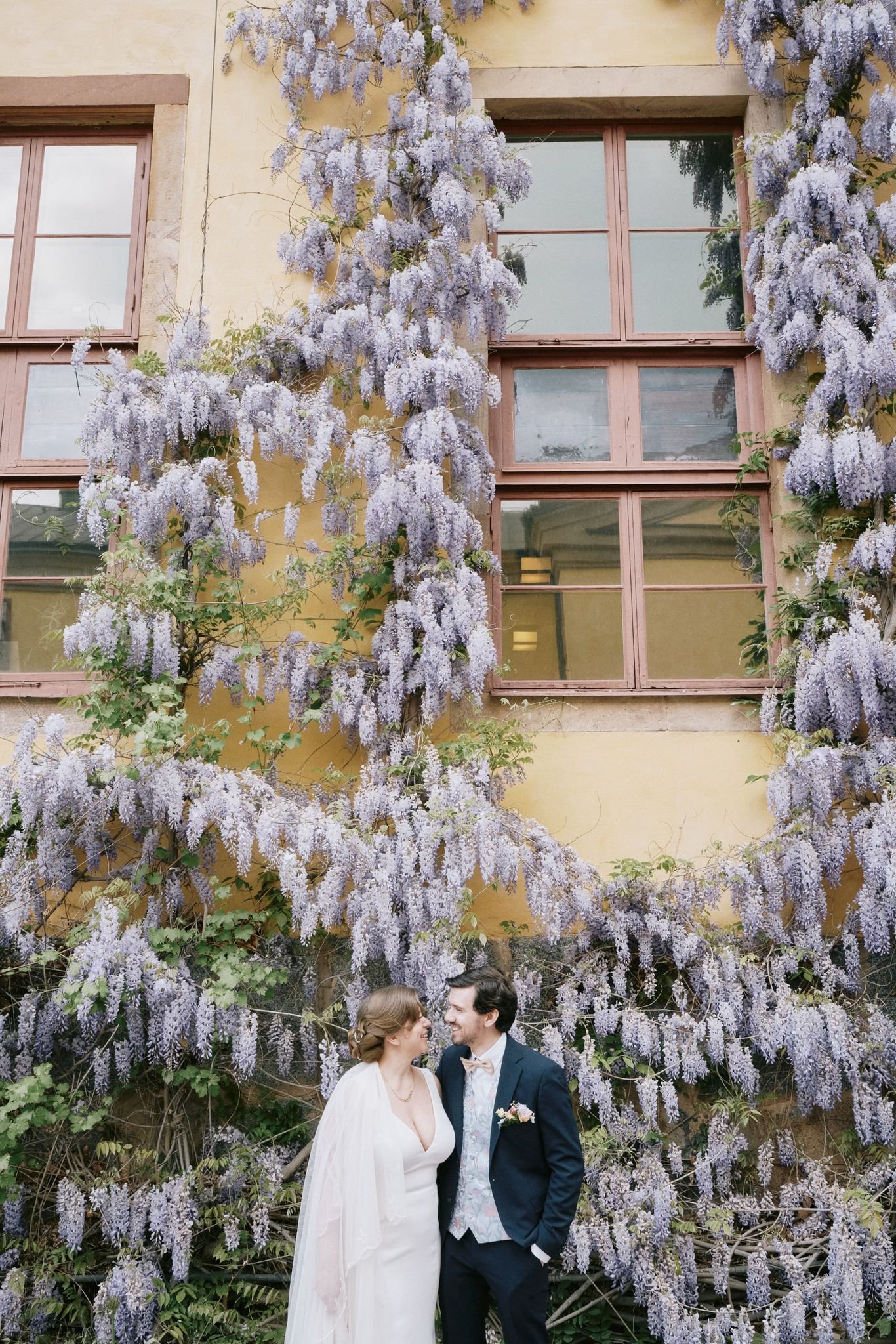 Ett par i bröllopskläder står framför en vägg täckt av lila blommande växter och ett gulaktigt byggnadsvägg.