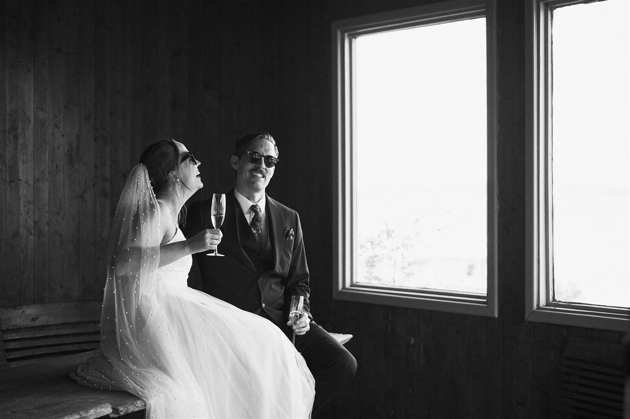 Ett brudpar sitter tillsammans vid ett fönster inomhus med dryck i handen