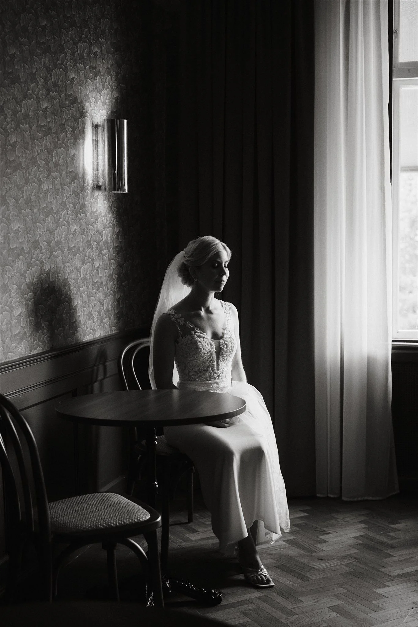 En svartvit bild av en brud i vardagsrummet som sitter på en stol, klädd i vigselklänning, nära ett fönster med vita gardiner.