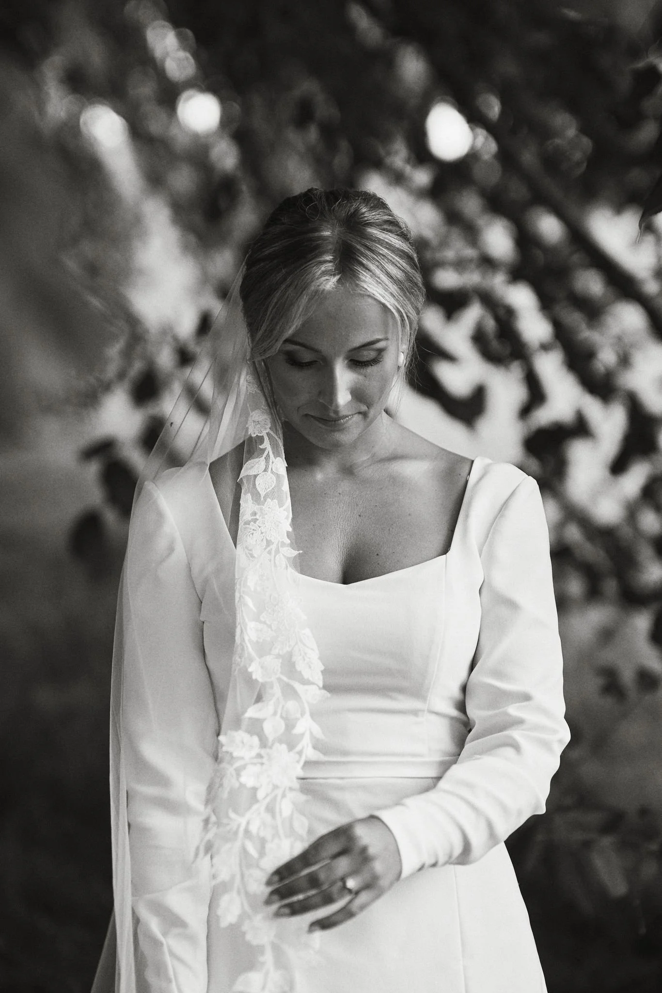 En kvinna i bröllopsklänning som tittar ner, med en blombukett i handen, och en blommig slöja på huvudet, svartvitt foto.