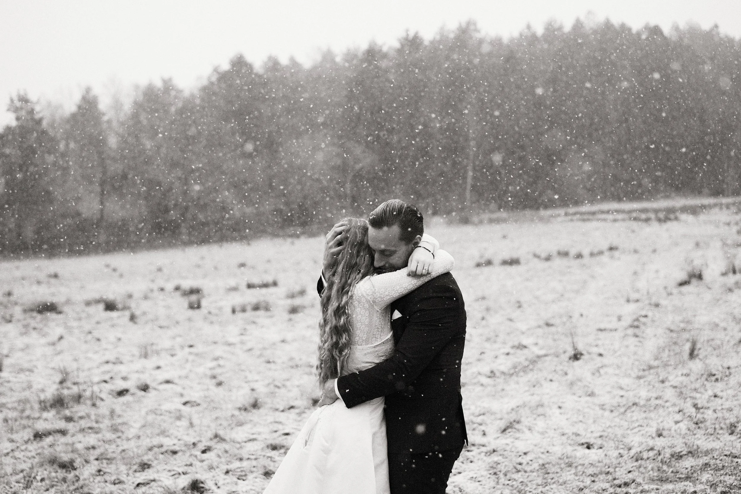 Ett brudpar omfamnar varandra i snöfall i ett öppet vinterlandskap