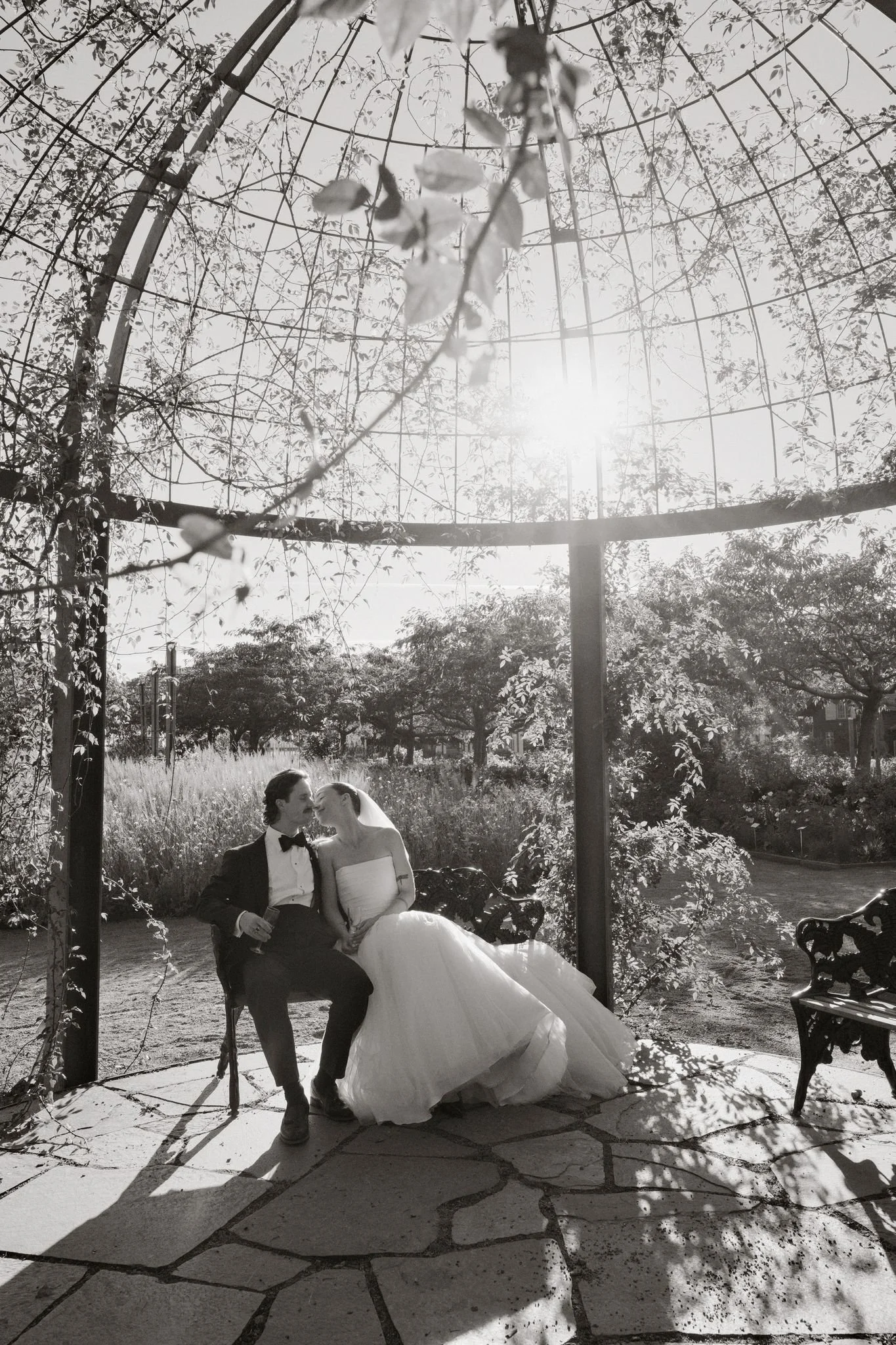 Ett brudpar sitter på en bänk under ett träd i en park, fotat i svartvitt, med solen i bakgrunden.