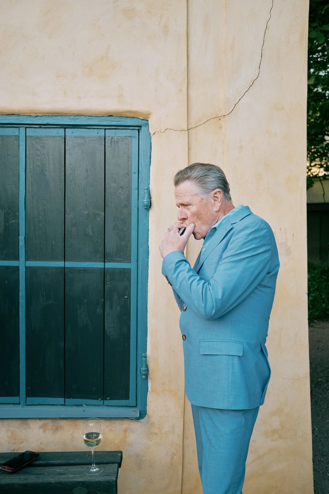 En äldre man i ljusblå kostym står utanför en gul vägg med sprickor, röker en cigarett och har en drink på ett bord framför sig.