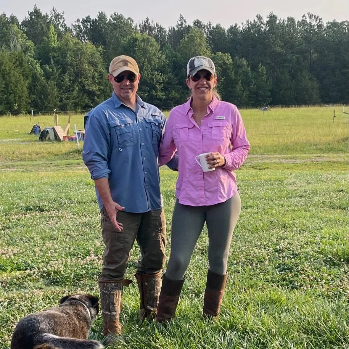 A man and woman standing in a grassy field during daytime, smiling at the camera. The man is wearing a blue button-up shirt, camouflage hat, sunglasses, and camo boots. The woman is wearing a pink button-up shirt, gray leggings, brown chaps, sunglasses, and a tan cap, holding a white mug. There is a dog lying on the grass in the foreground and trees in the background.