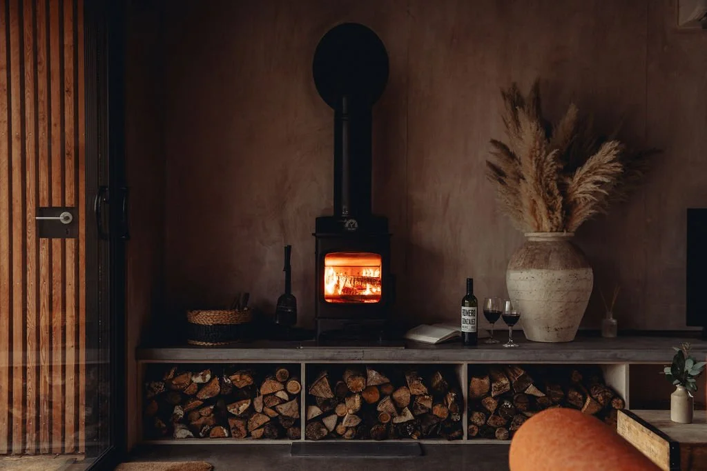 A cozy interior with a burning wood stove, a large vase with pampas grass, a bottle of wine, two glasses, a book, and stacked firewood below.