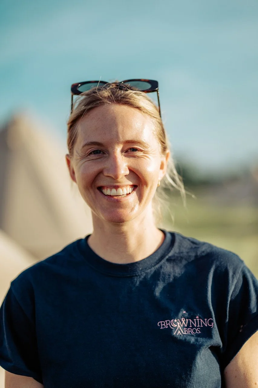 A smiling person wearing sunglasses on their head and a navy blue T-shirt with "Browning Bros" written on it, outdoors against a blurry background.
