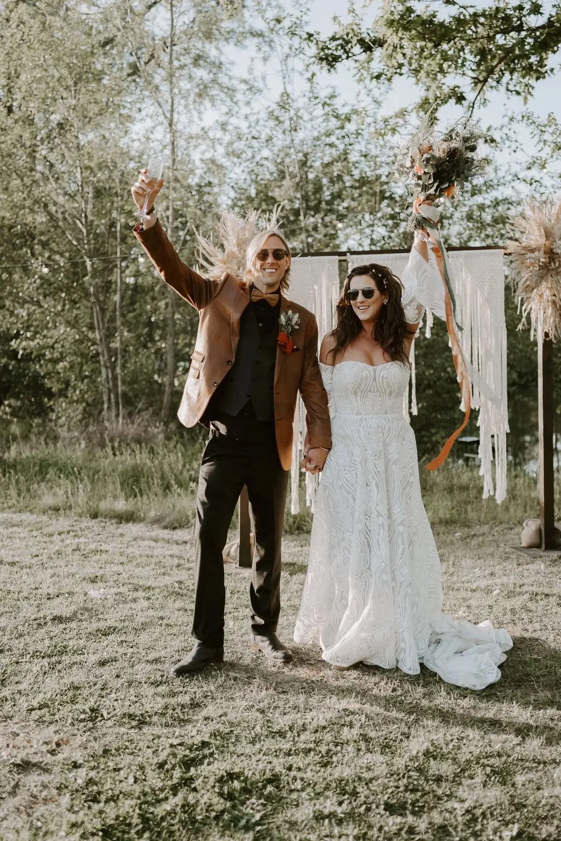 Bride and groom celebrating outdoors with sunglasses and wedding attire in natural setting.