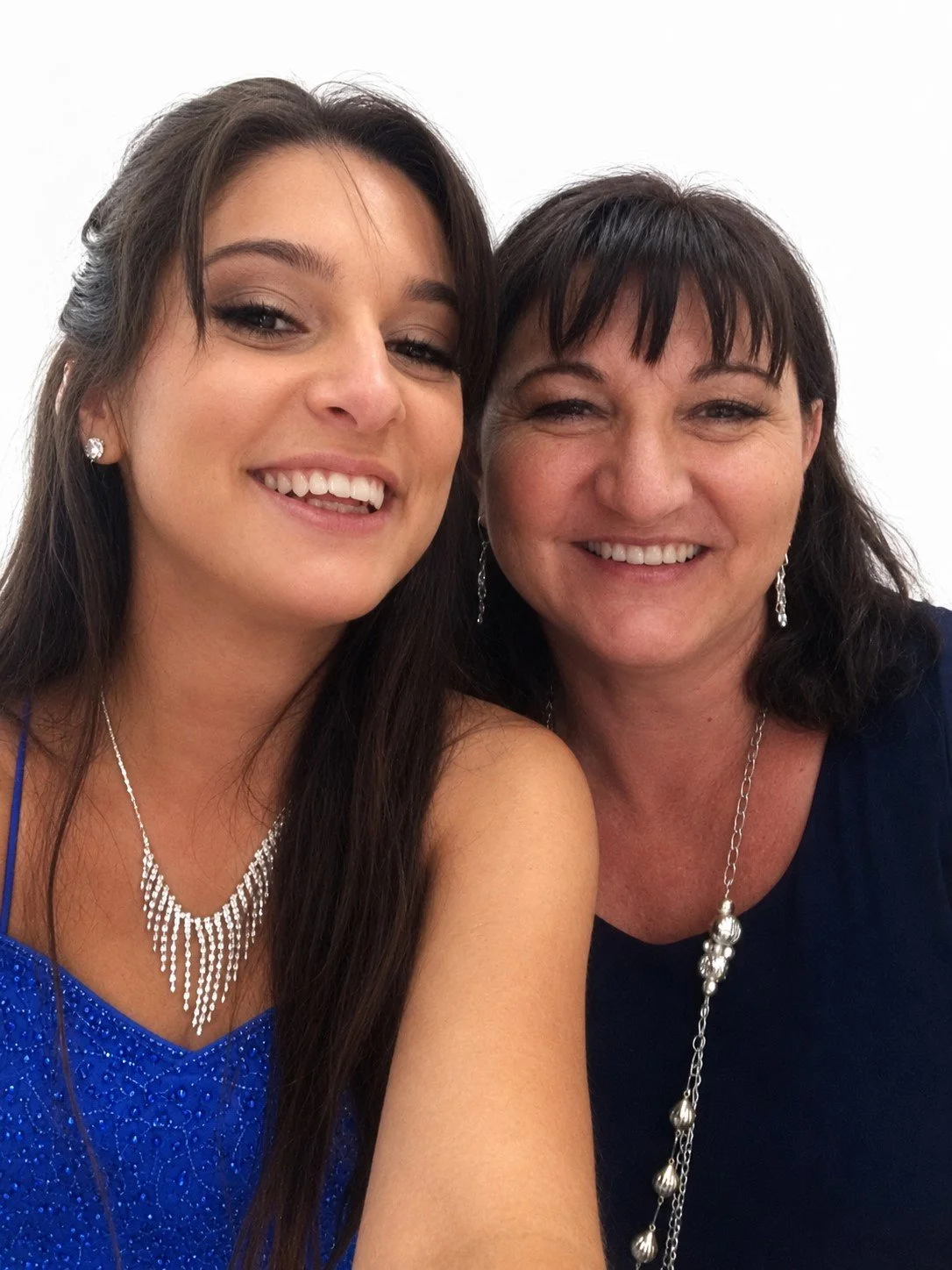 Two women smiling and taking a selfie against a plain white background. The younger woman on the left has long brown hair, is wearing a blue dress with sparkles, and jewelry including earrings and a necklace. The older woman on the right has short dark hair, is wearing a dark top with jewelry including earrings and a long necklace.