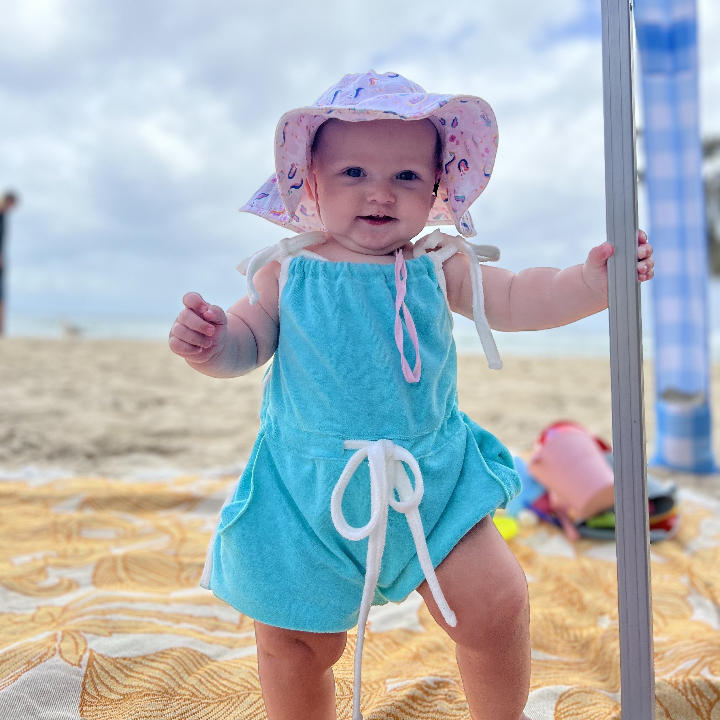 A smiling baby girl in a blue dress and pink sun hat standing on a beach, holding onto a metal pole.