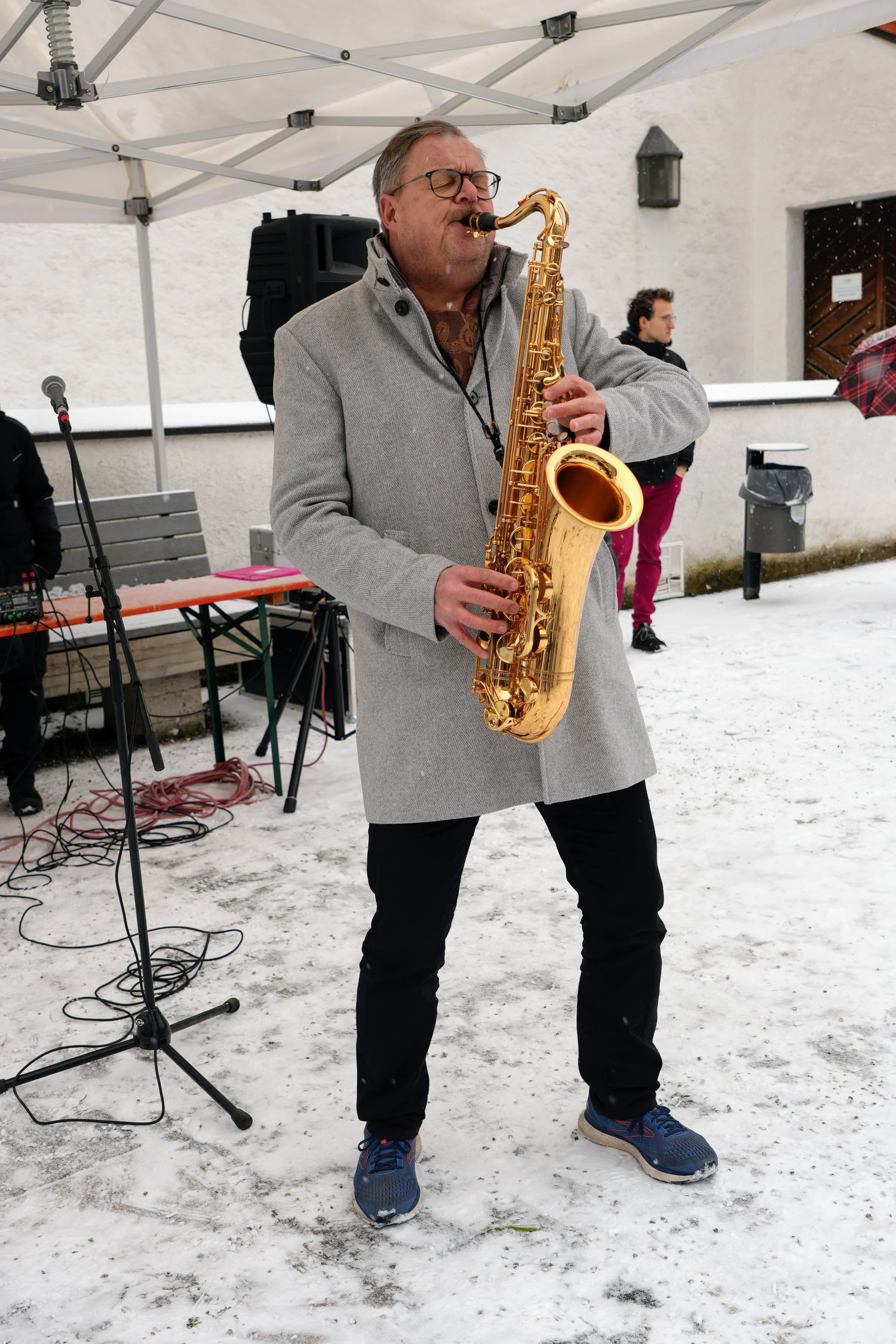 Ein Mann spielt auf einem Goldenen Saxophon im Freien bei winterlichem Wetter, mit Schneeboden und anderen Menschen im Hintergrund.