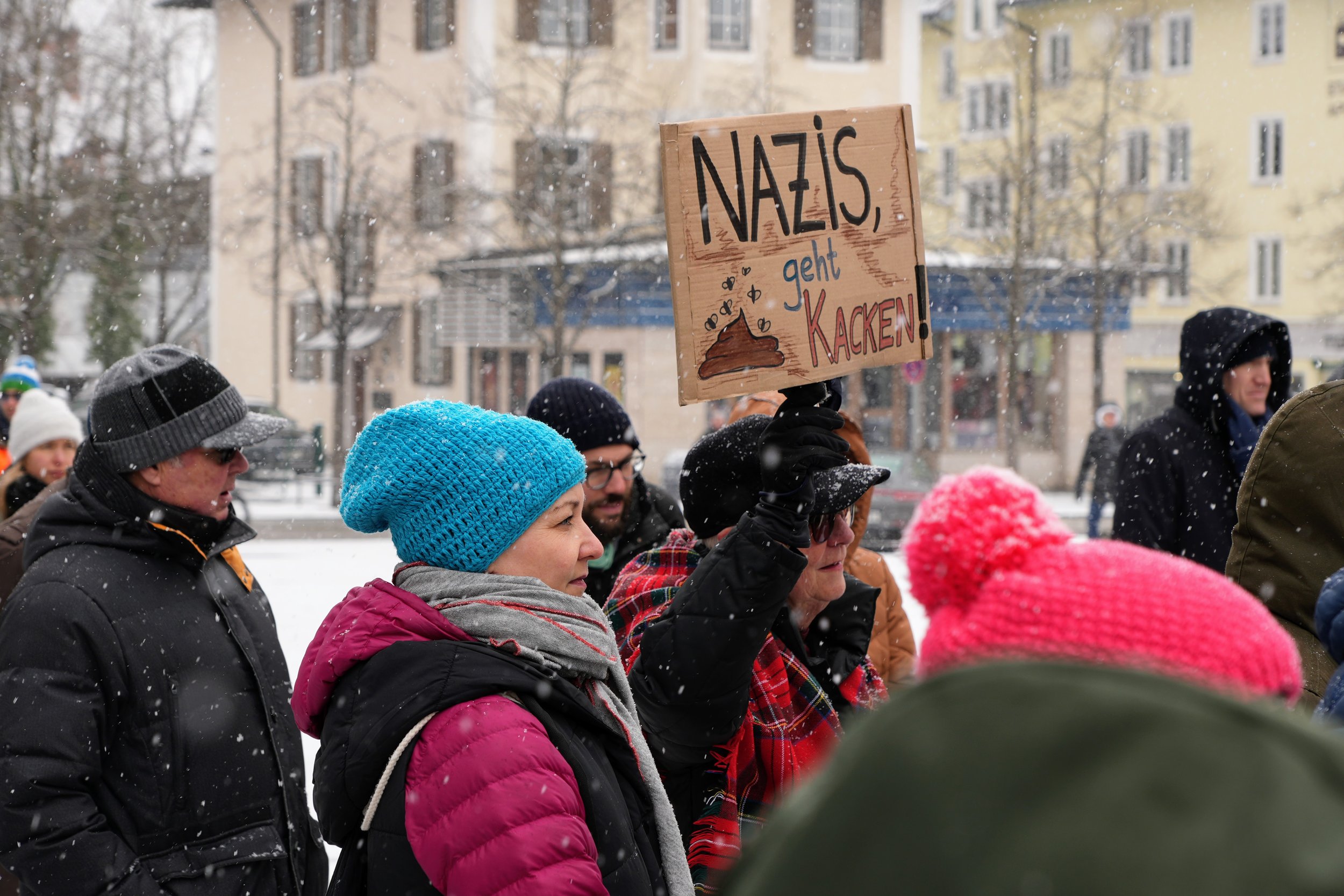 Menschen bei einer Demonstration im Schnee, eine Frau hält ein Schild mit der Aufschrift 'Nazis geht Kacken'.