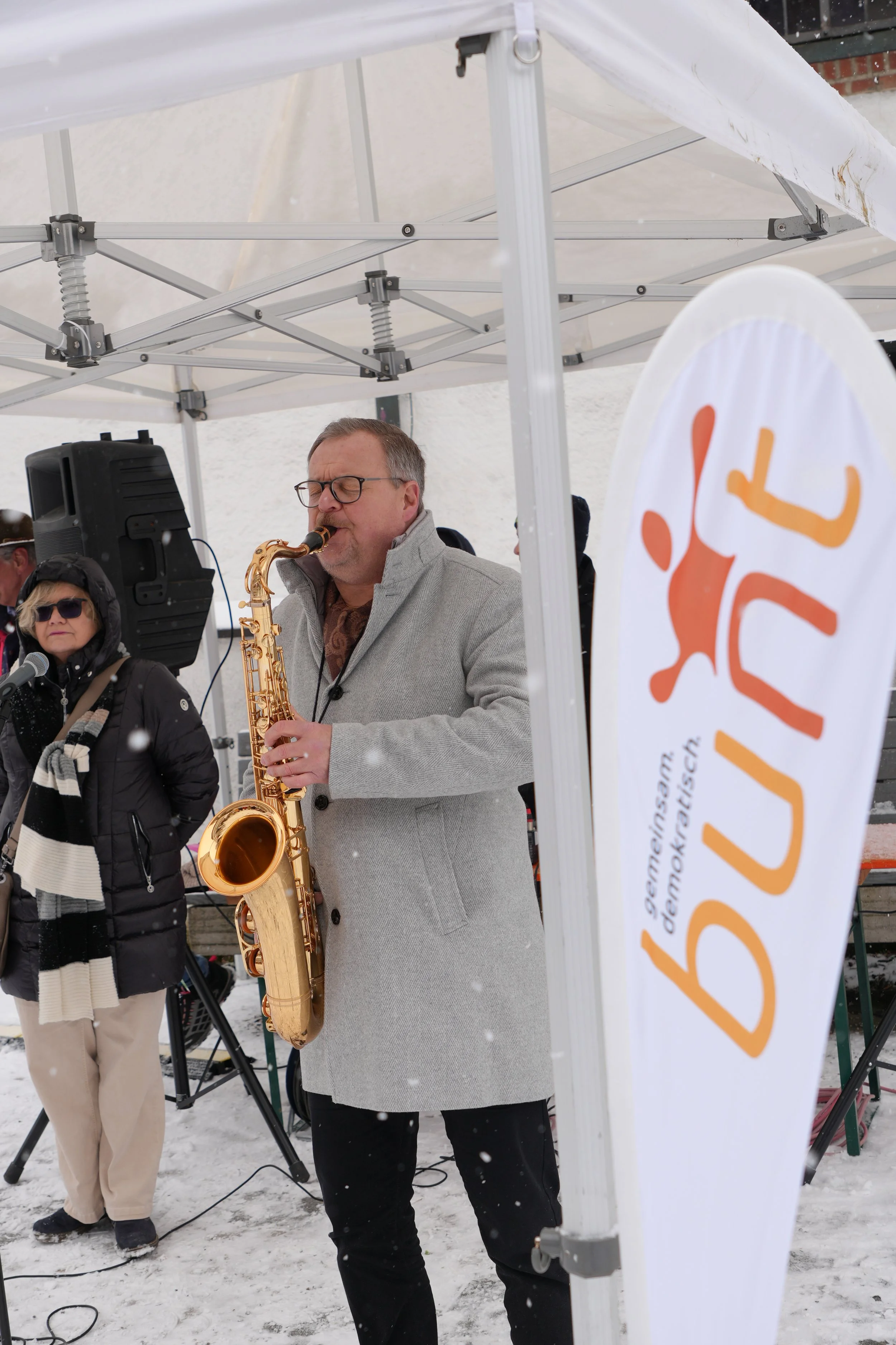 Ein Mann spielt Saxophon während eines Events im Freien im Schnee, mit einer Frau im Hintergrund und einem weißen Zelt.