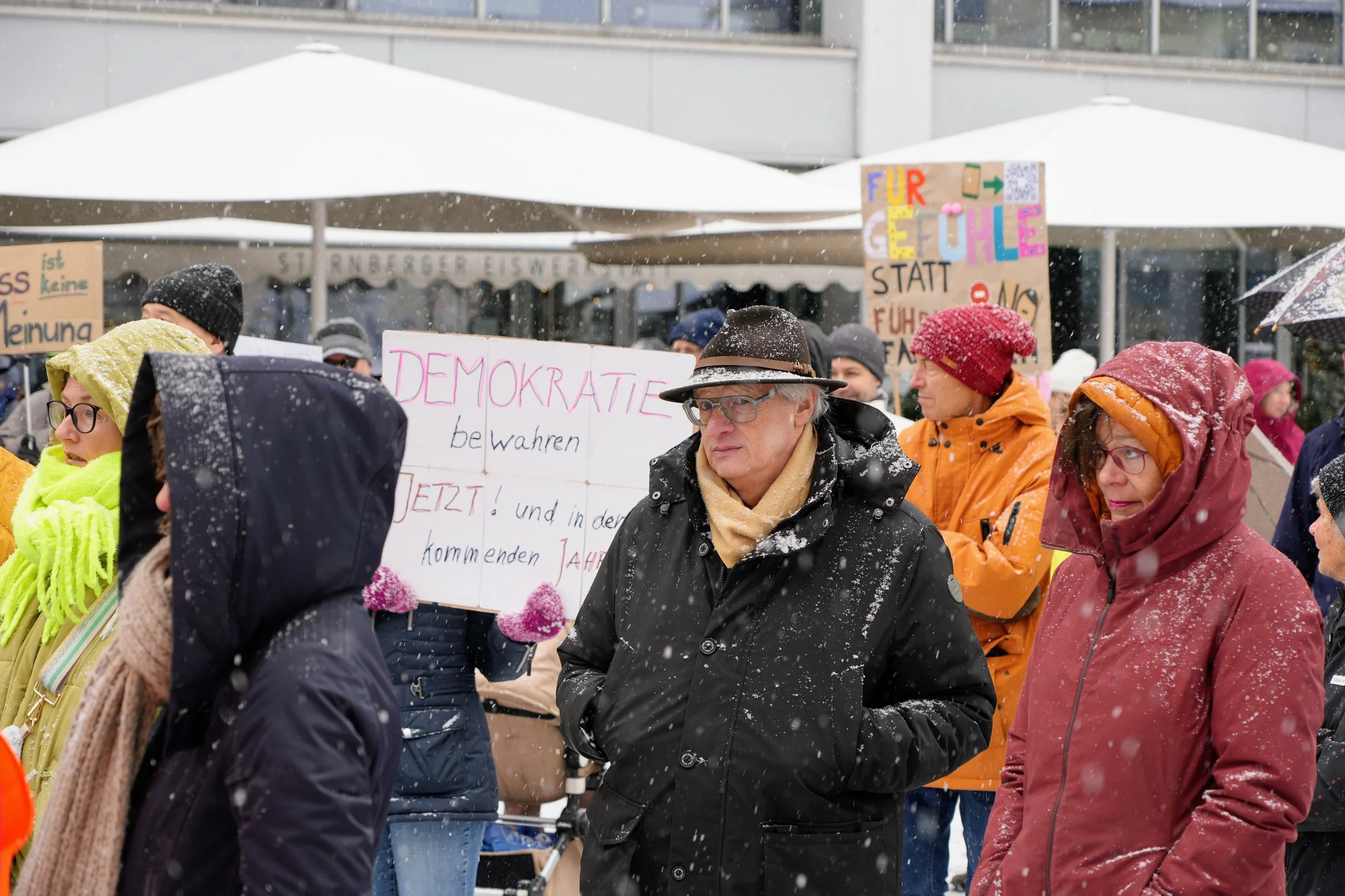 Menschen bei einer Demonstration im Schneefall, halten Schilder mit politischen Botschaften, some wearing winter clothing, gläserne Bauten im Hintergrund.