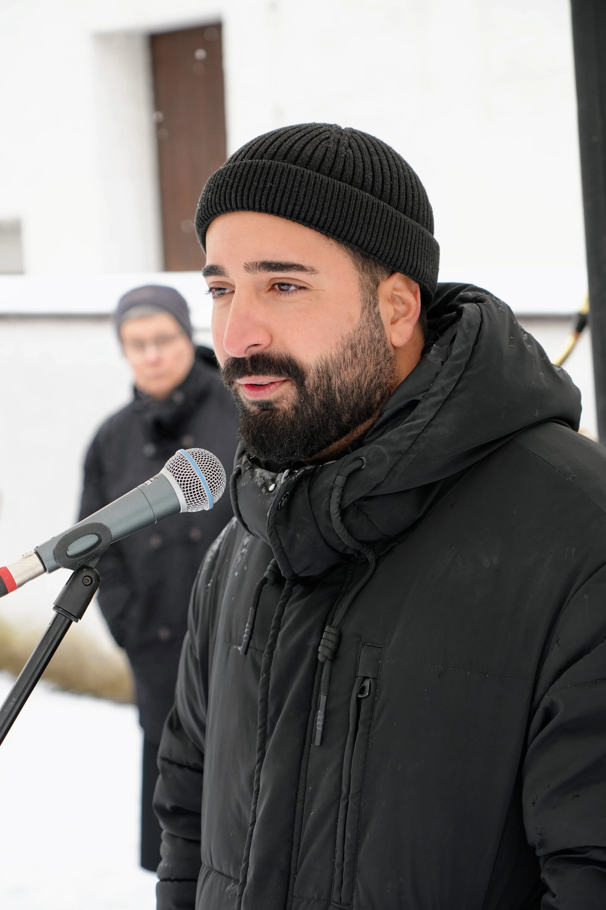 Mann mit schwarzem Wintermantel und schwarzen Mütze spricht vor einem Mikrofon, im Hintergrund eine weitere Person.