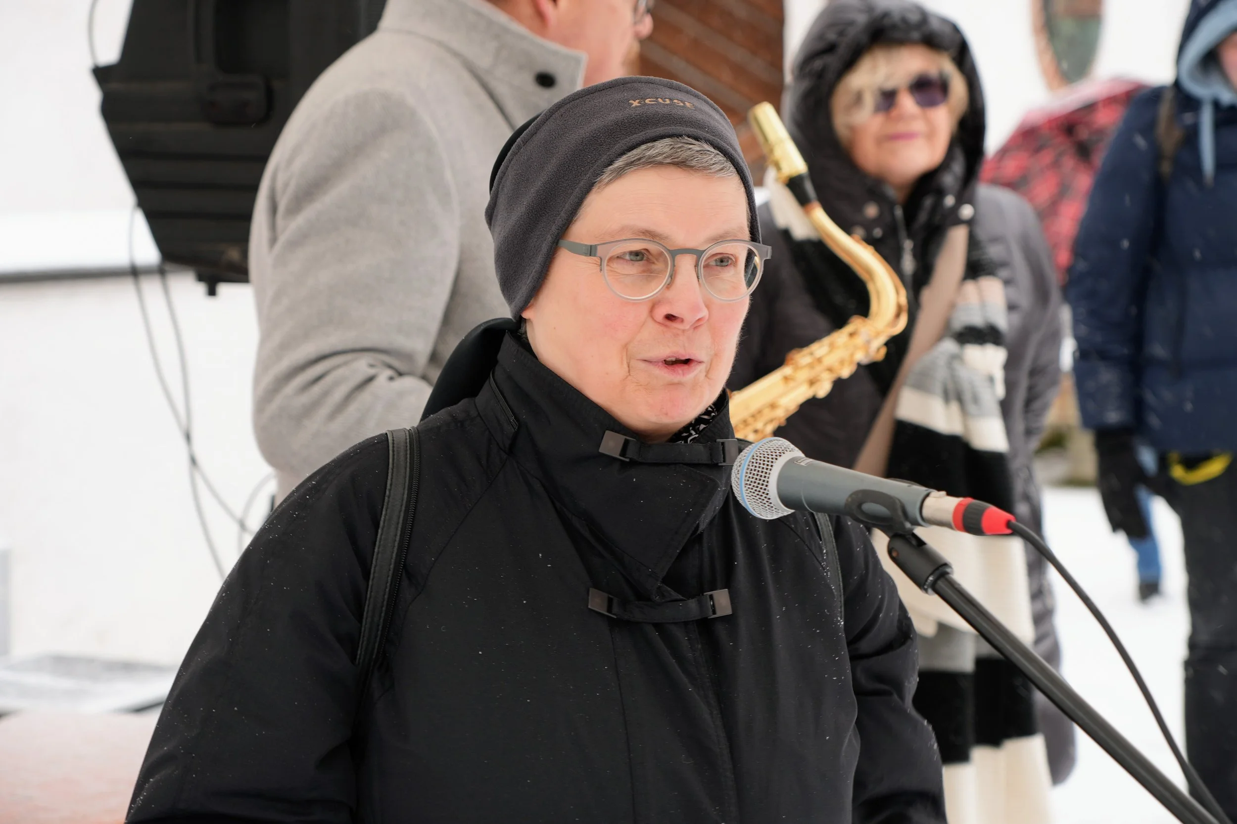 Eine Frau trägt eine schwarze Jacke, eine schwarze Mütze und eine Brille, spricht in ein Mikrofon, im Hintergrund sind mehrere Personen, darunter eine Person mit einer Saxophon, bei kaltem Wetter im Freien.