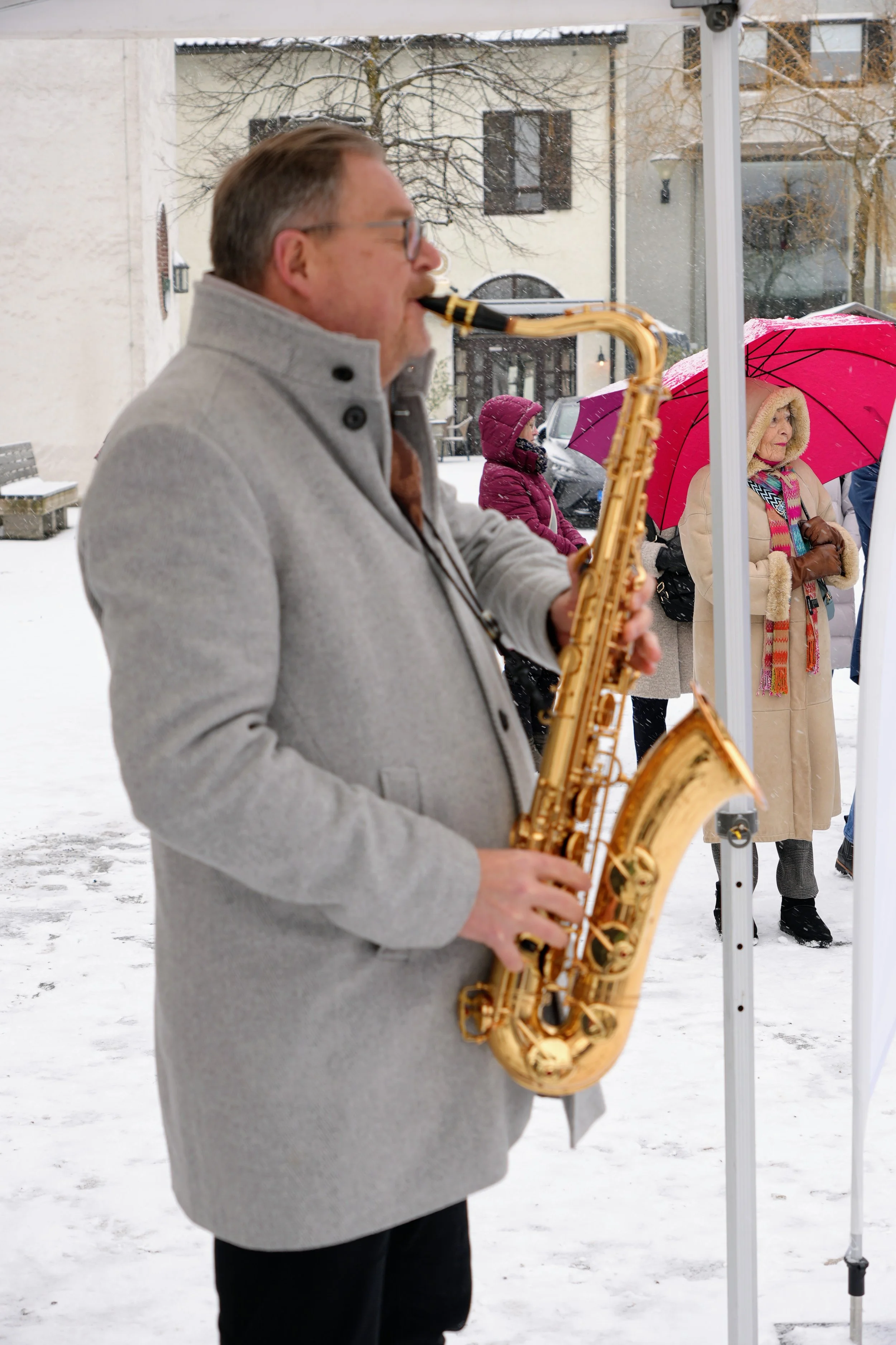 Ein Mann spielt Saxophon im Schnee bei einem Winterevent, im Hintergrund Frauen mit Regenschirmen und Winterkleidung.