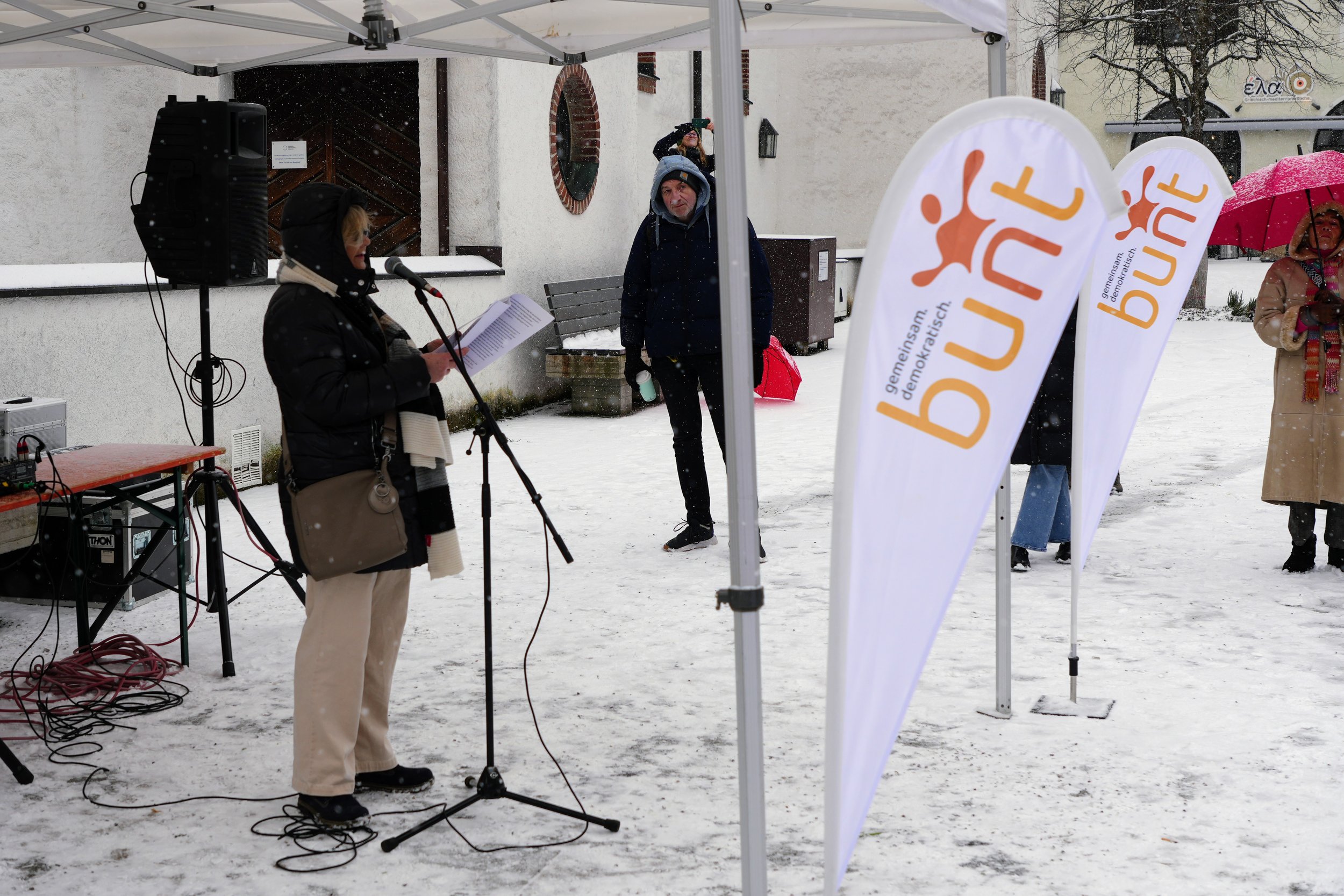 Eine Frau liest bei einer Veranstaltung im Freien im Schnee, während andere Personen mit Regenschirmen im Hintergrund stehen. Es gibt zwei Banner mit der Aufschrift „Bündnis“ und eine Bühne mit Mikrofon und Lautsprecher.