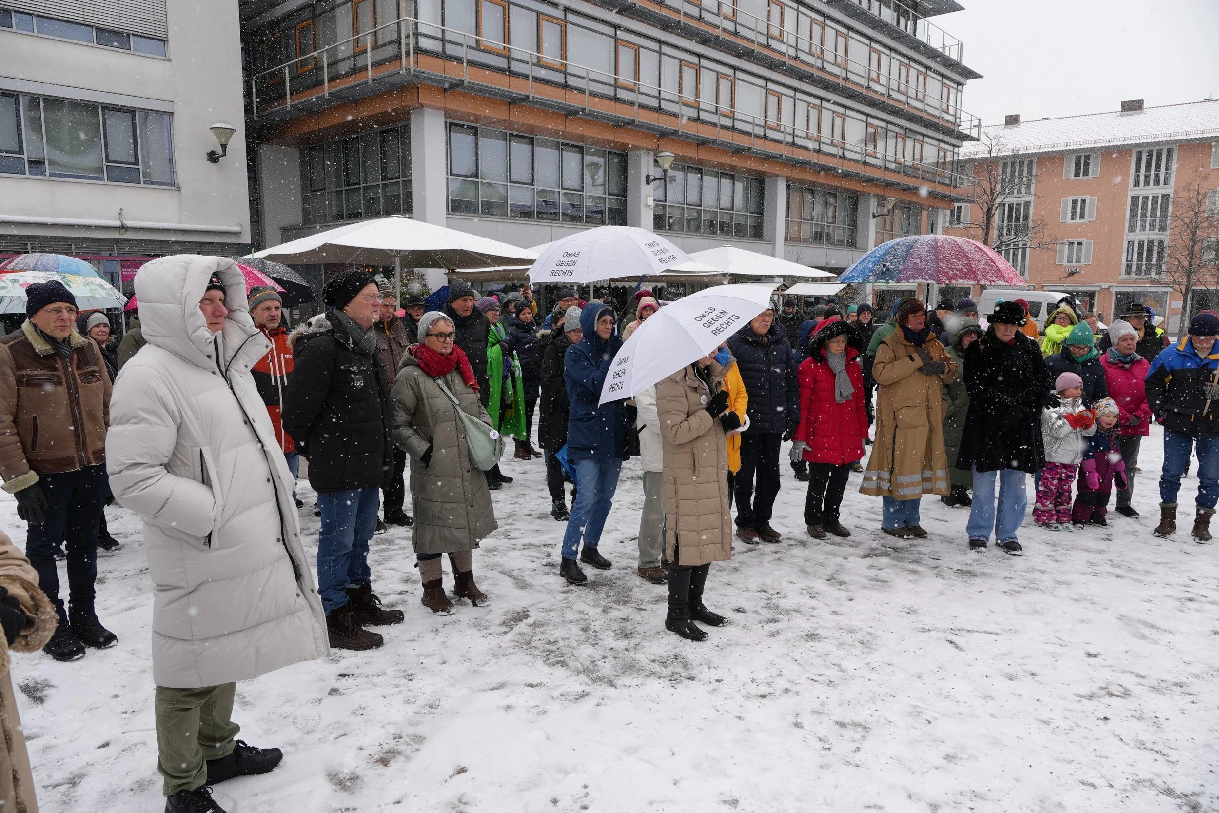 Personen versammeln sich im Schneefall bei einer Demonstration im Freien, einige halten Schirme mit Aufdrucken, die gegen rechts sind, in einer urbanen Umgebung mit mehrstöckigen Gebäuden im Hintergrund.