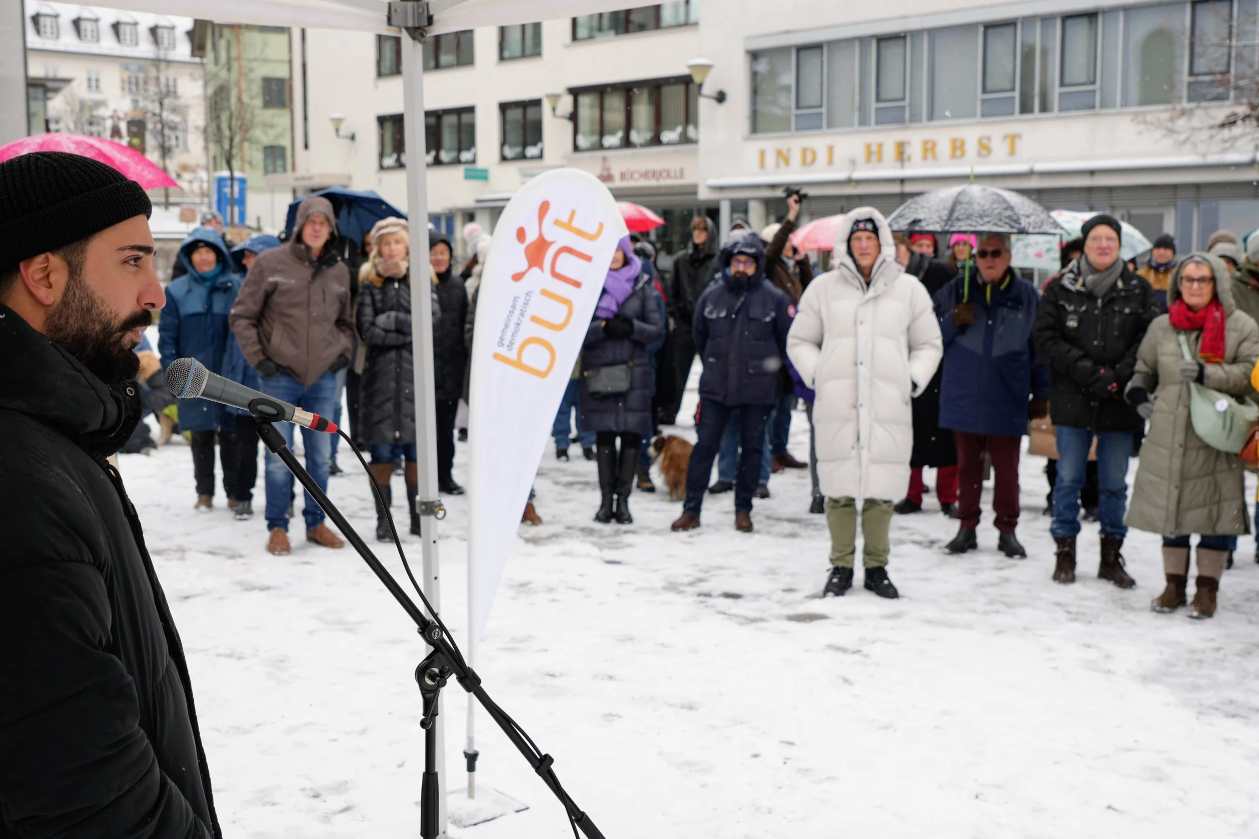 Ein Mann spricht mit einem Mikrofon vor einer Menschenmenge, die im Schnee steht, mit Regenschirmen und Winterkleidung, bei einem öffentlichen Anlass im Winter.