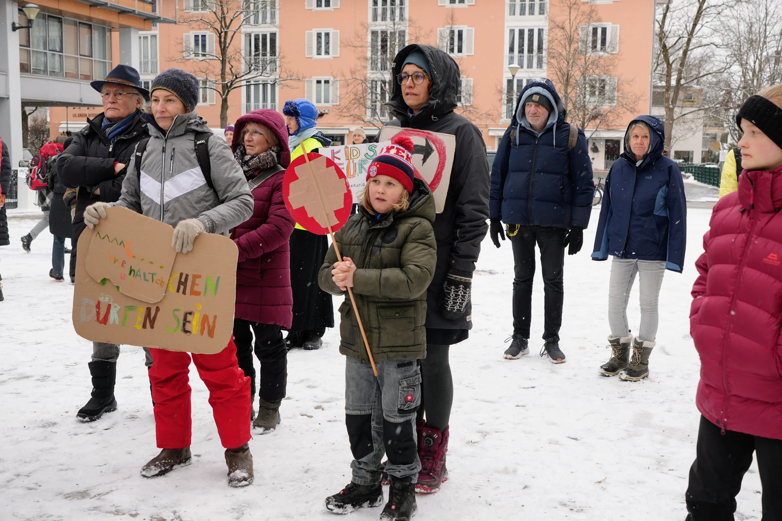 Menschen nehmen an einer Demonstration im Schnee teil und halten Schilder mit Botschaften wie 'Kinderschuhe dürfen sein'.