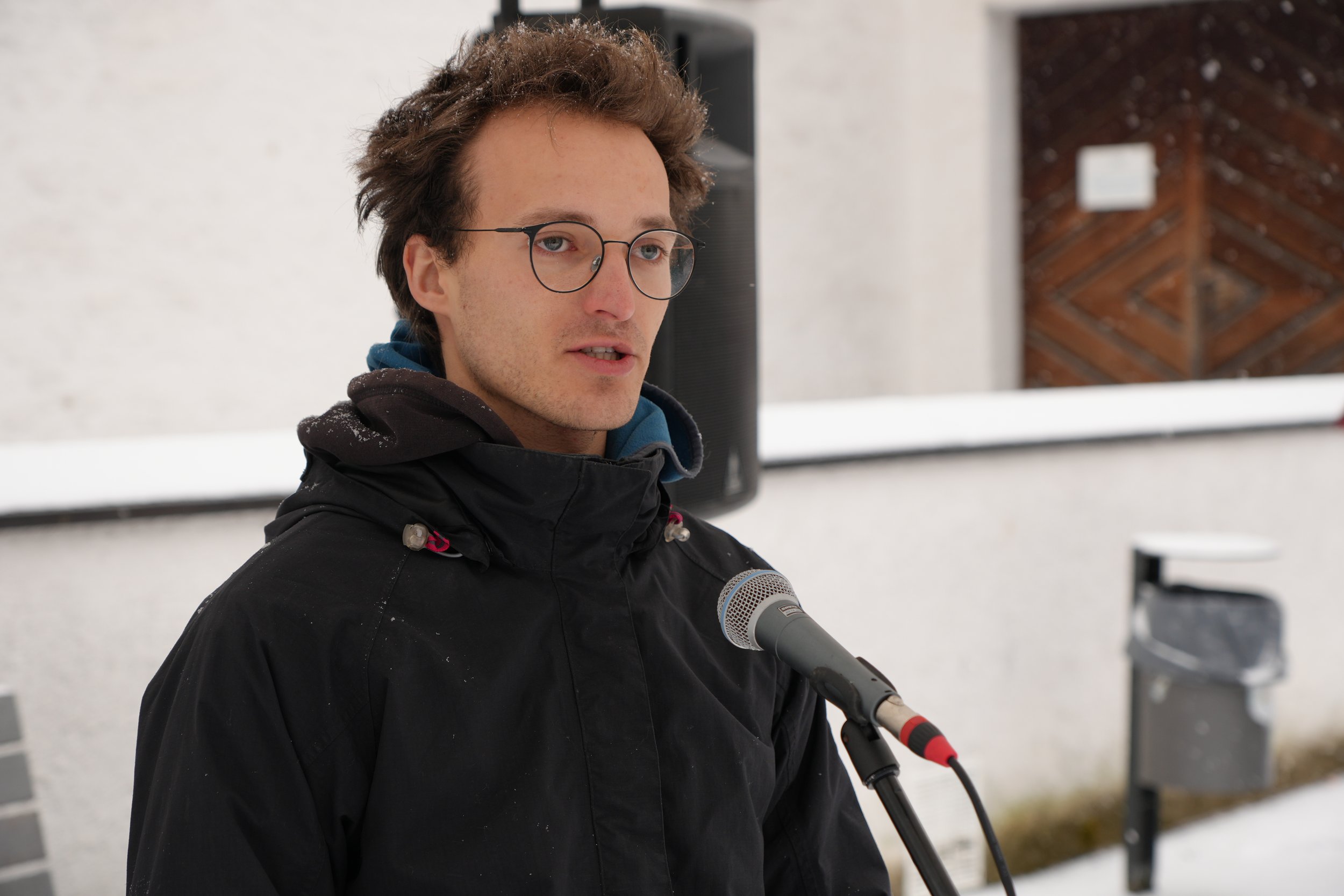 Junger Mann spricht in einem Mikrofon im Freien, trägt eine schwarze Jacke, Winterkleidung, Brille und hat braunes, lockiges Haar, umgeben von Schnee.