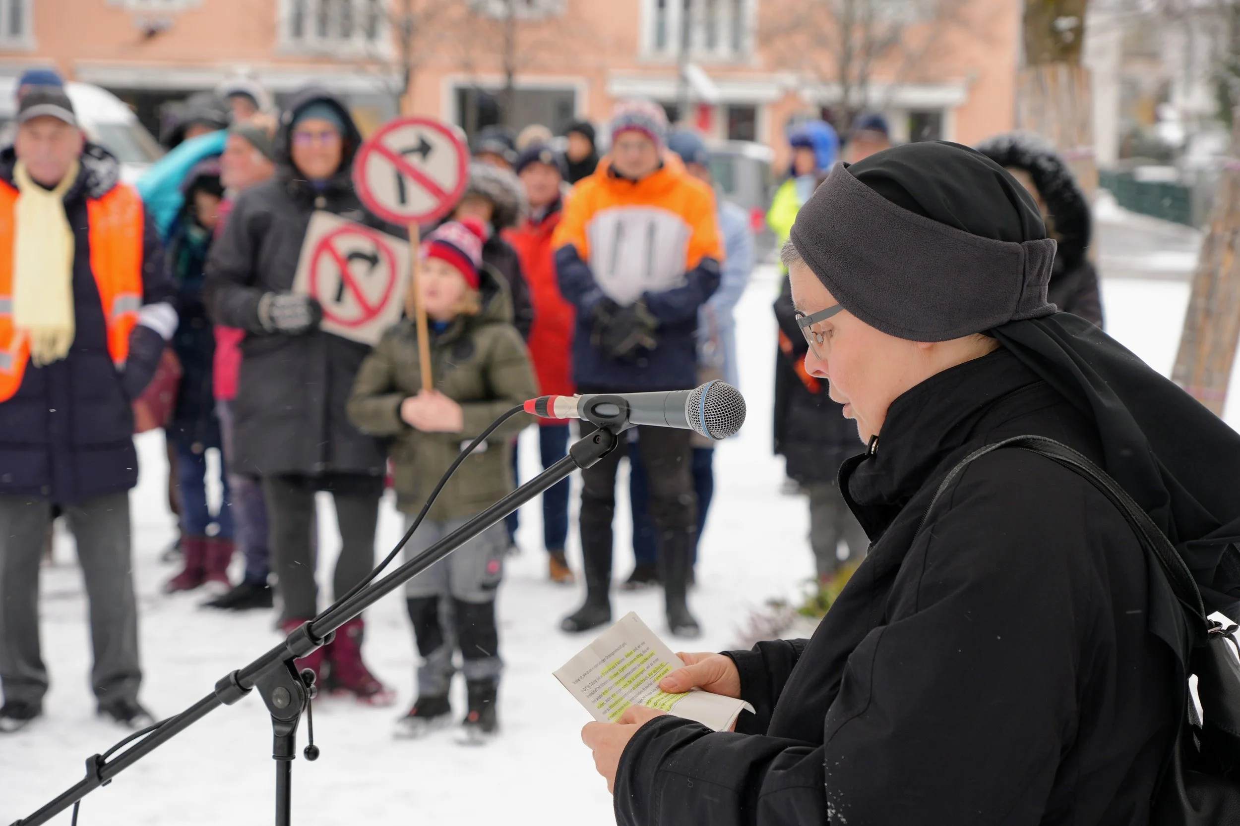Eine Person liest vor einem Mikrofon bei einer Protestkundgebung im Freien im Schnee, während im Hintergrund eine Gruppe von Menschen mit Schildern steht.