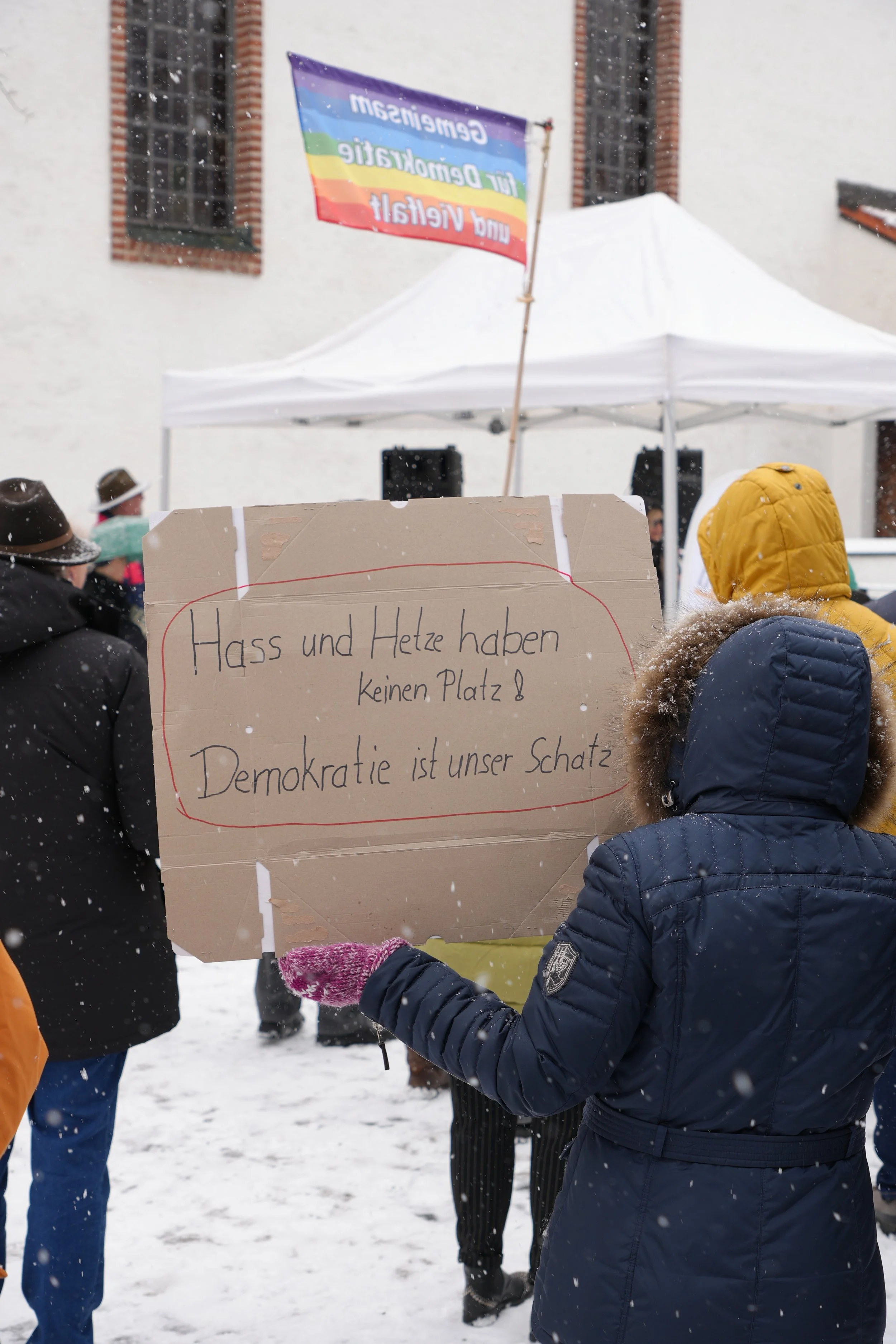 Ein Demonstrant hält ein Plakat mit der Aufschrift: 'Hass und Hetze haben keinen Platz! Demokratie ist unser Schatz'. Im Hintergrund sind Menschen mit Winterkleidung beim Protest in Schneewetter. Es gibt ein Zelt und eine Regenbogenflagge.