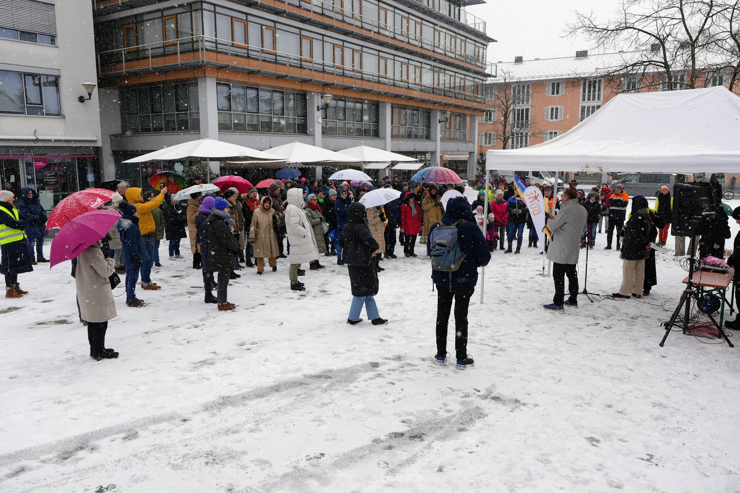 Menschen versammeln sich bei einer Veranstaltung im Schnee, viele halten Regenschirme, in einem Stadtplatz mit Gebäuden im Hintergrund