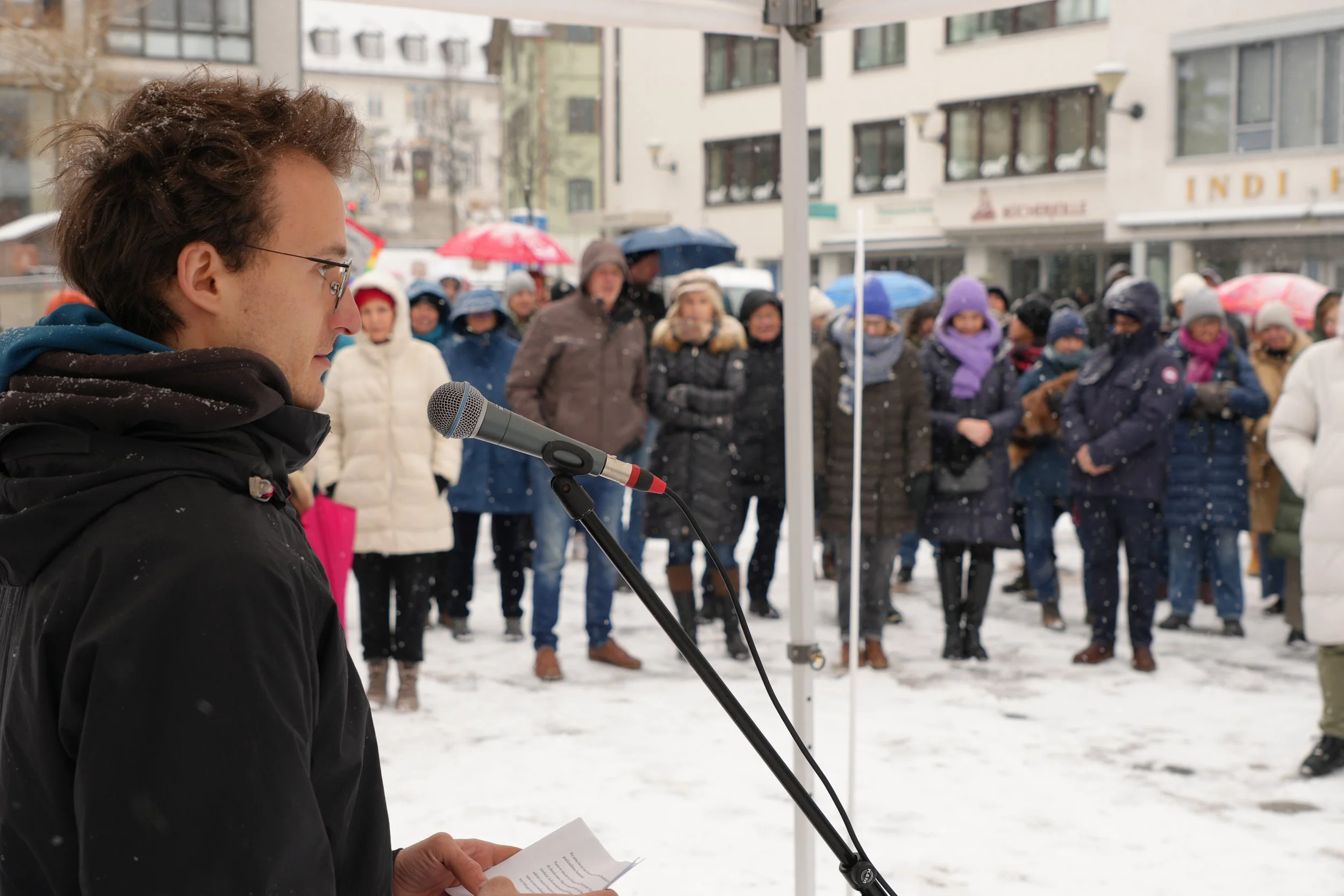Ein junger Mann liest bei einer Demonstration im Schnee, während eine Menschenmenge im Hintergrund zuhört, im Hintergrund befindet sich ein Stadtzentrum mit Gebäuden und Geschäften.