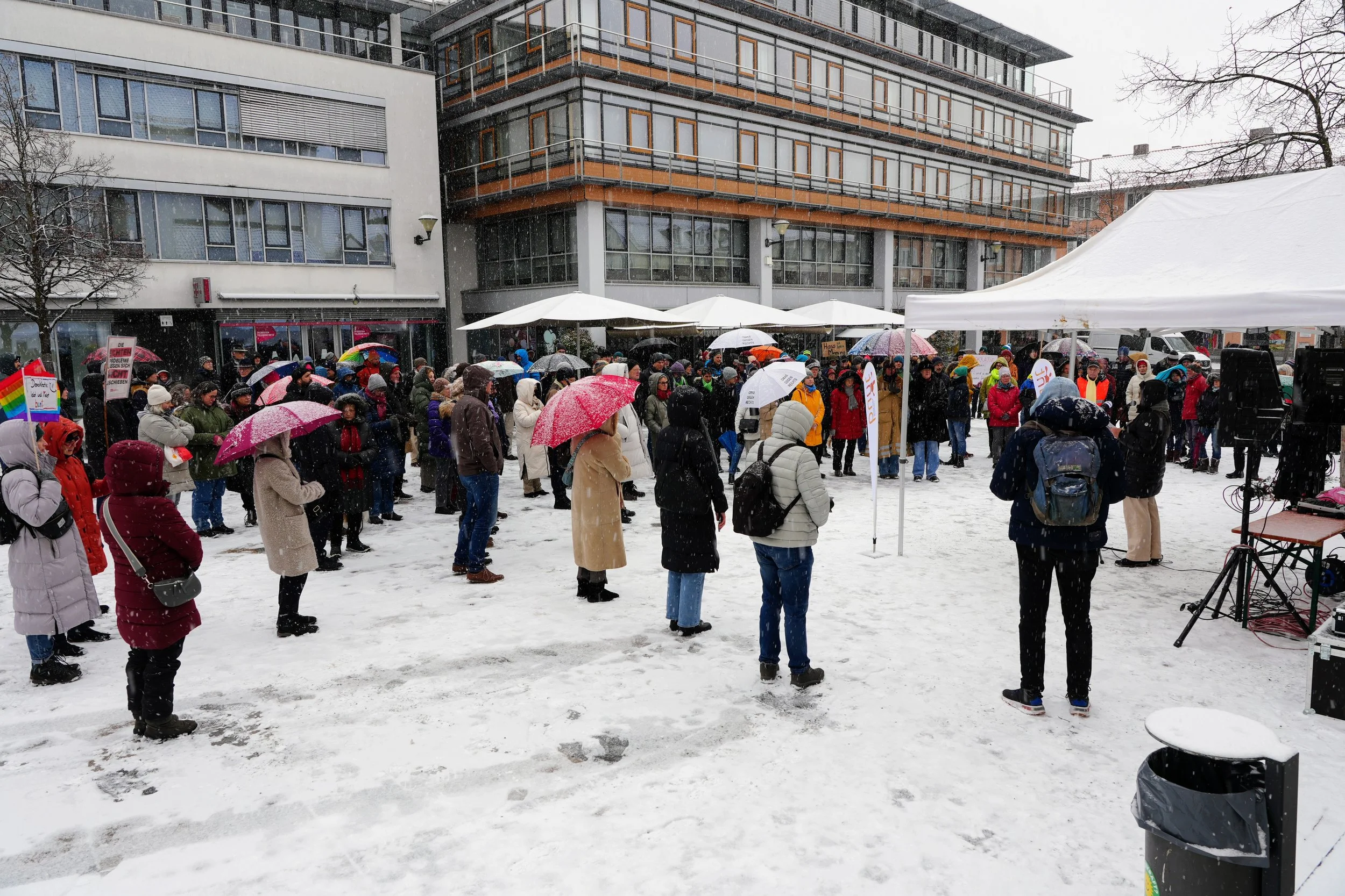 Menschen versammeln sich bei einer Demonstration im Schnee, einige mit Regen- oder Schneeschirm, in einer urbanen Umgebung mit modernen Gebäuden und Zelten im Hintergrund.