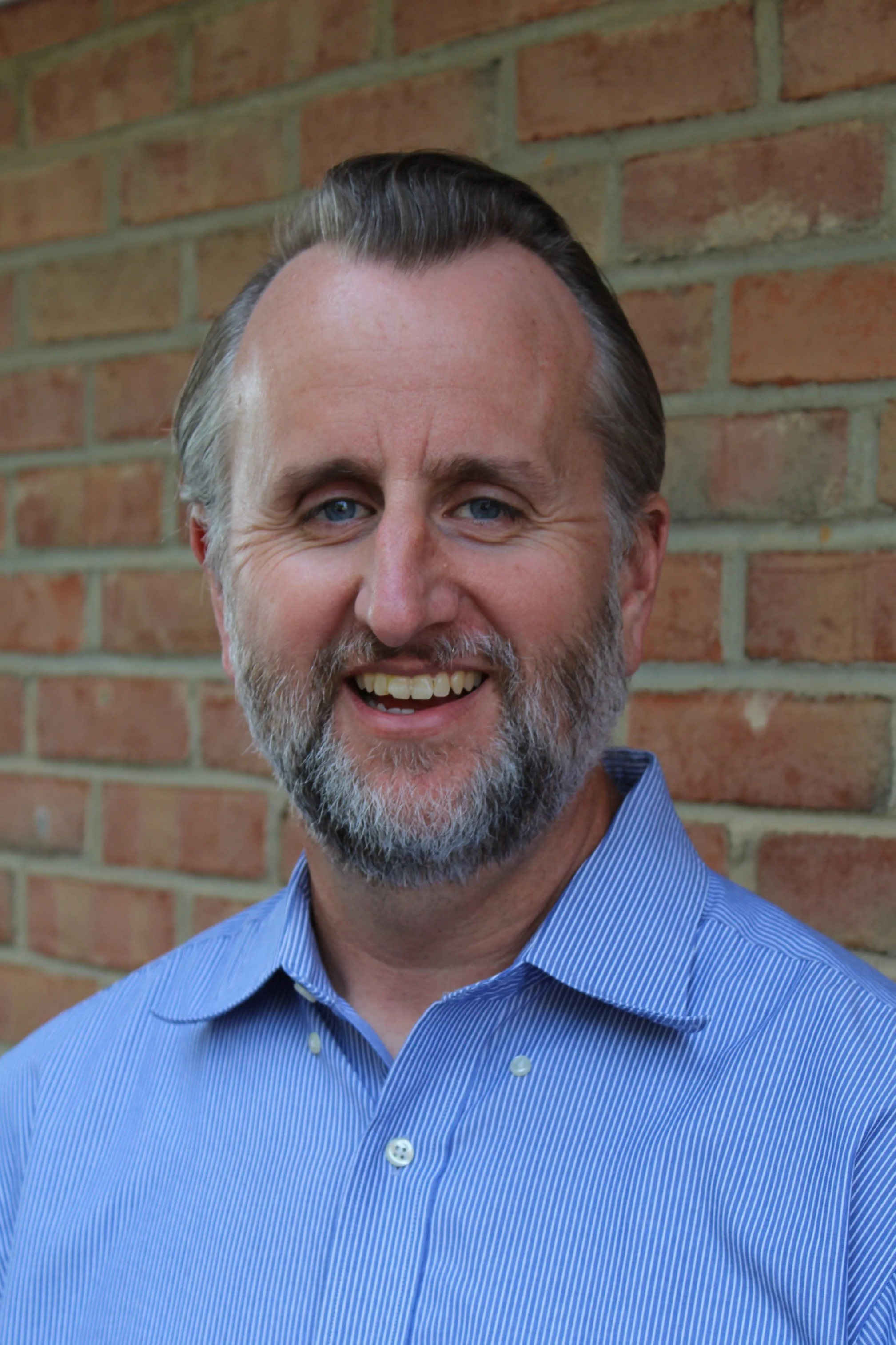 Outdoor headshot of a man with short gray hair and a beard, wearing a button up collared shirt.