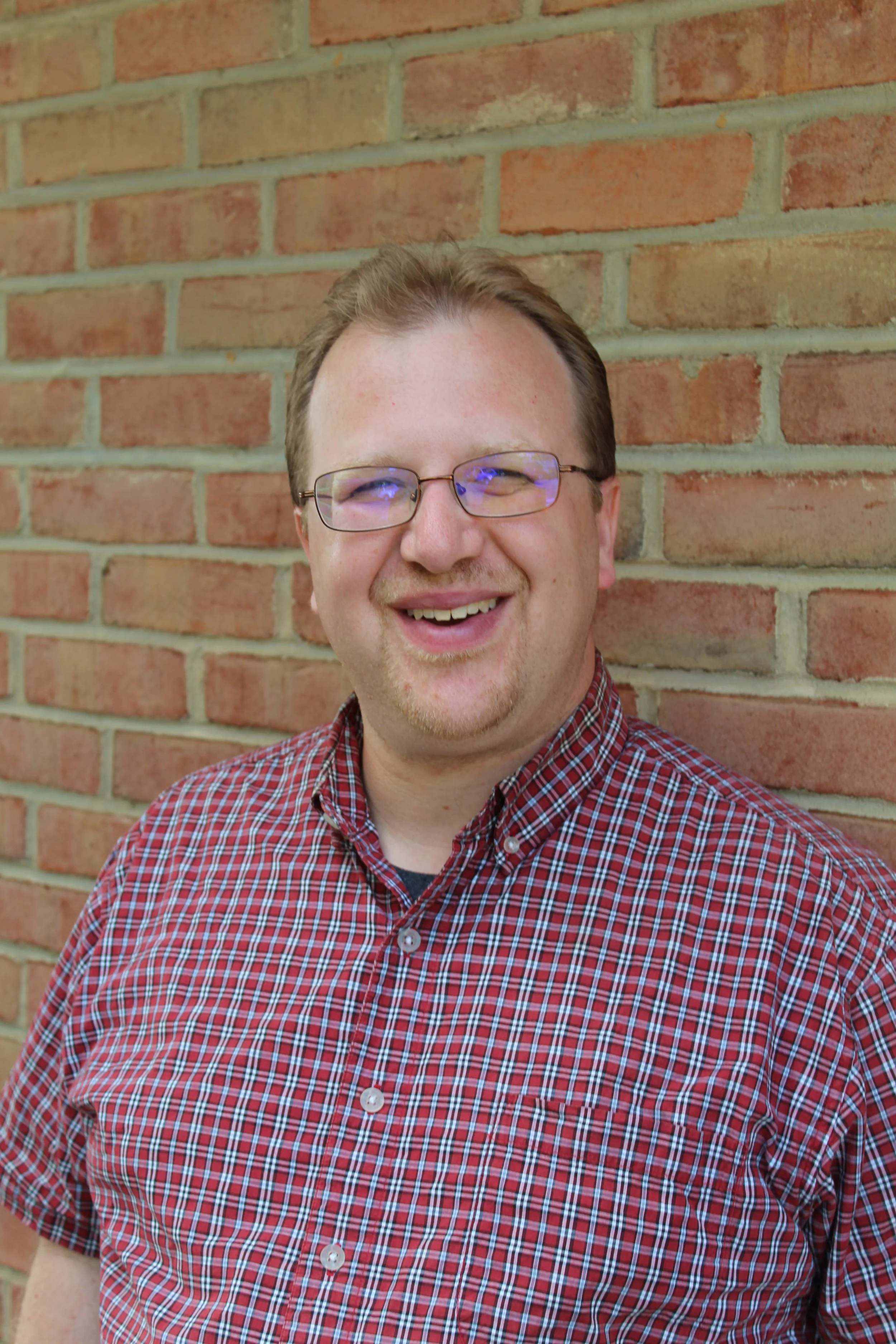 Headshot of Jay Trovato, a man with short blonde hair and glasses, smiling, wearing a red plaid collared button up shirt, standing in front of a brick wall.