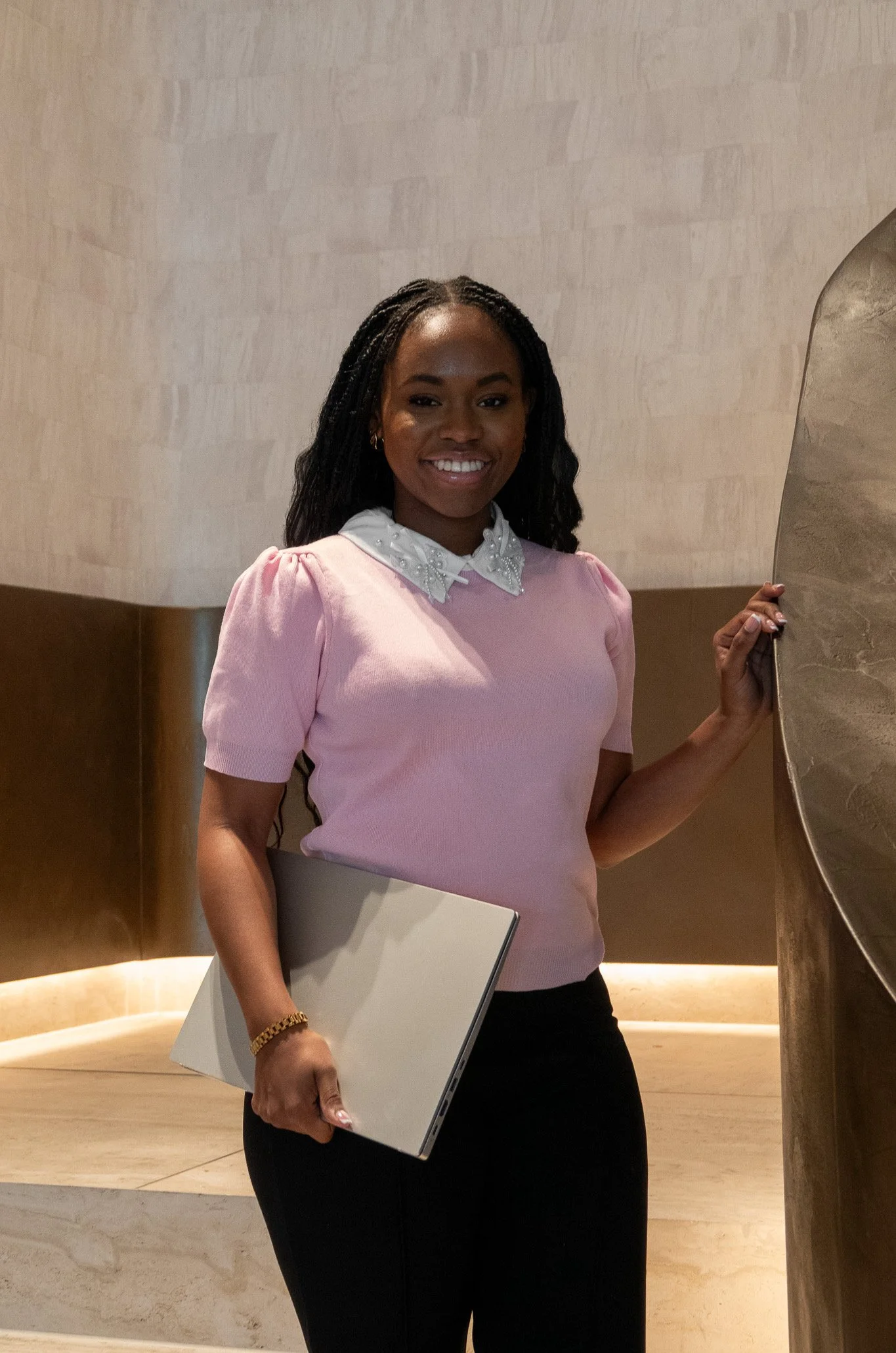 Vrouw met donkere huid, zwart haar, roze blouse met witte details, houdt een laptop en staat naast een grote gouden sculpture in een decoratief interieur.