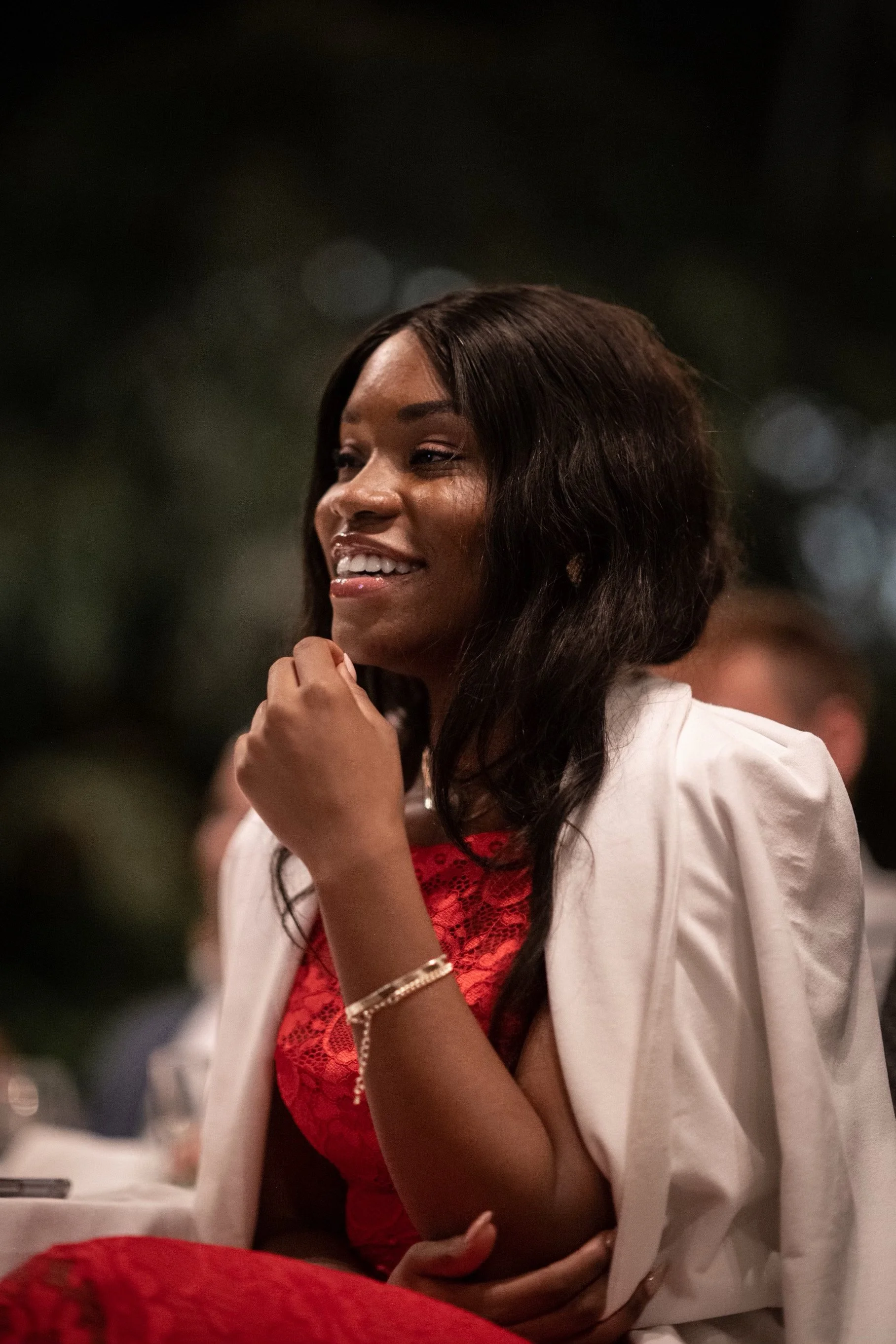 Een vrouw in een rode jurk en witte jas lacht en zit aan een tafel, met een wazige achtergrond.