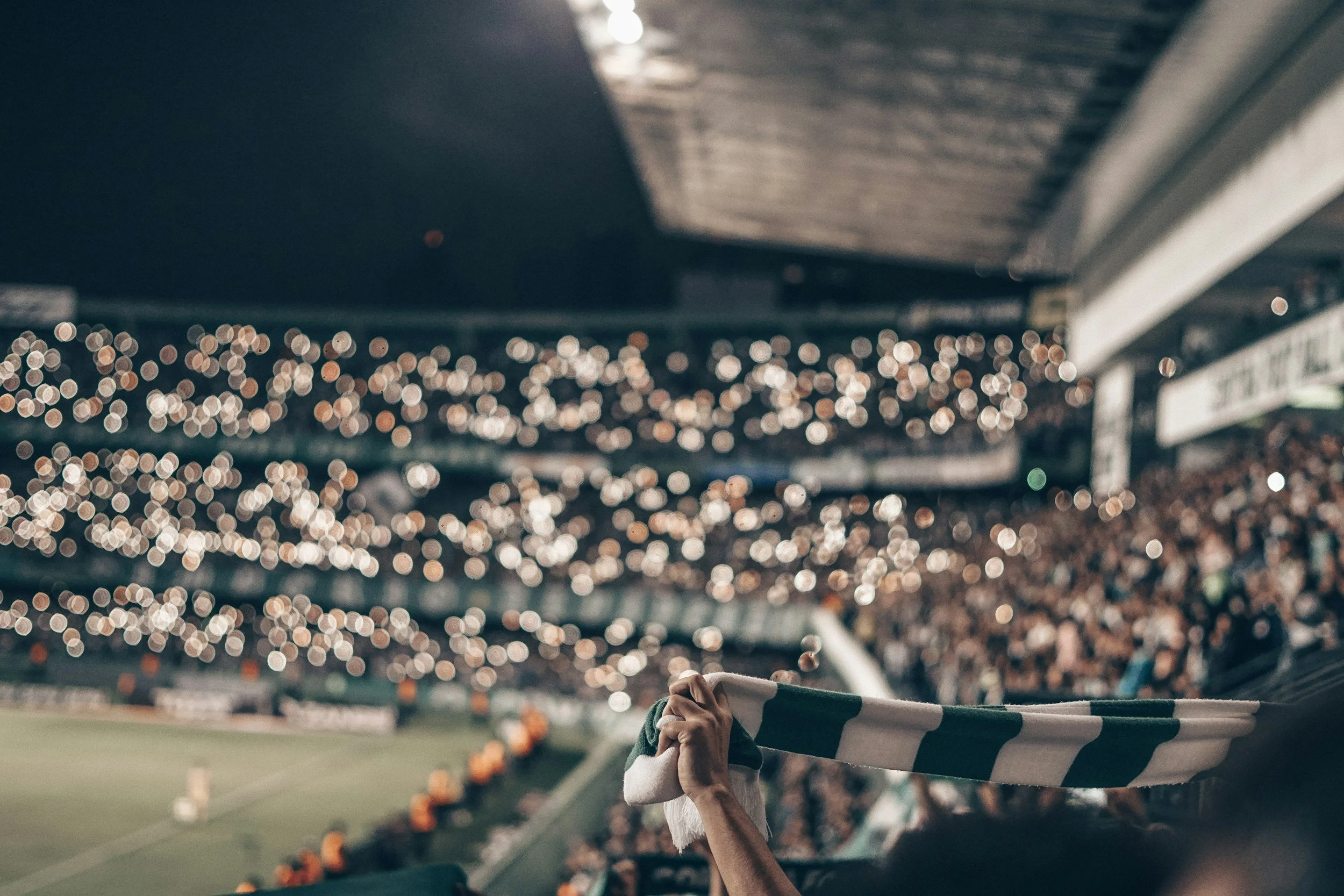 A person holding a scarf or cloth above their head inside a stadium during a night game, with blurred lights and spectators in the background.