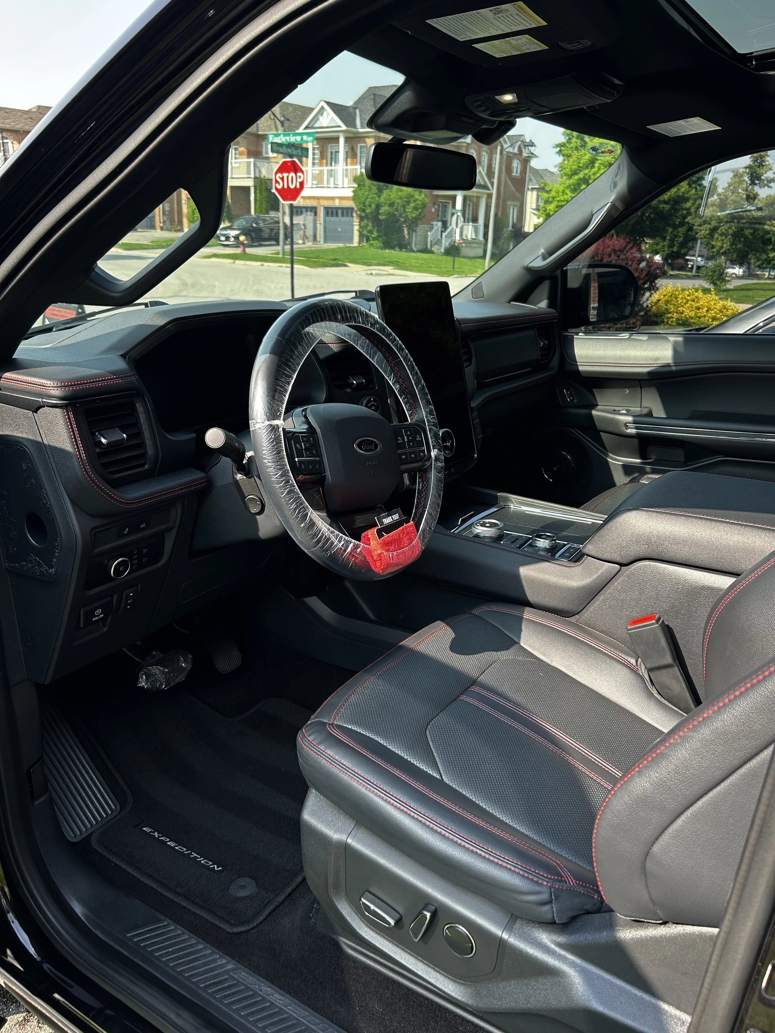 Inside view of a Ford vehicle with a wrapped steering wheel, black interior with red stitching, and a side view mirror showing modern houses and a stop sign outside.