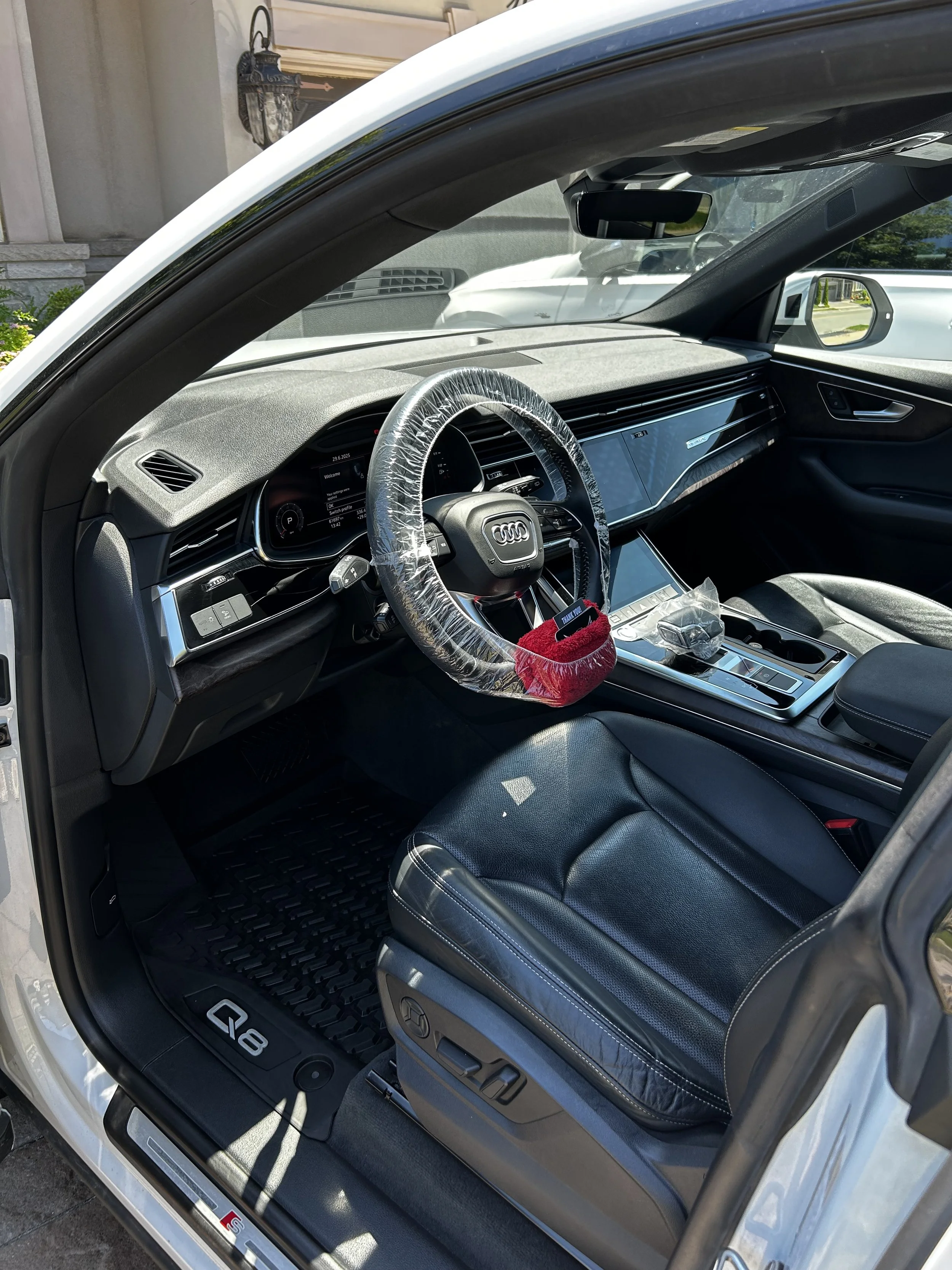 Interior of an Audi Q8 SUV showing the steering wheel, dashboard, gear shifter, and front seats, with a plastic steering wheel cover and various items on the center console.